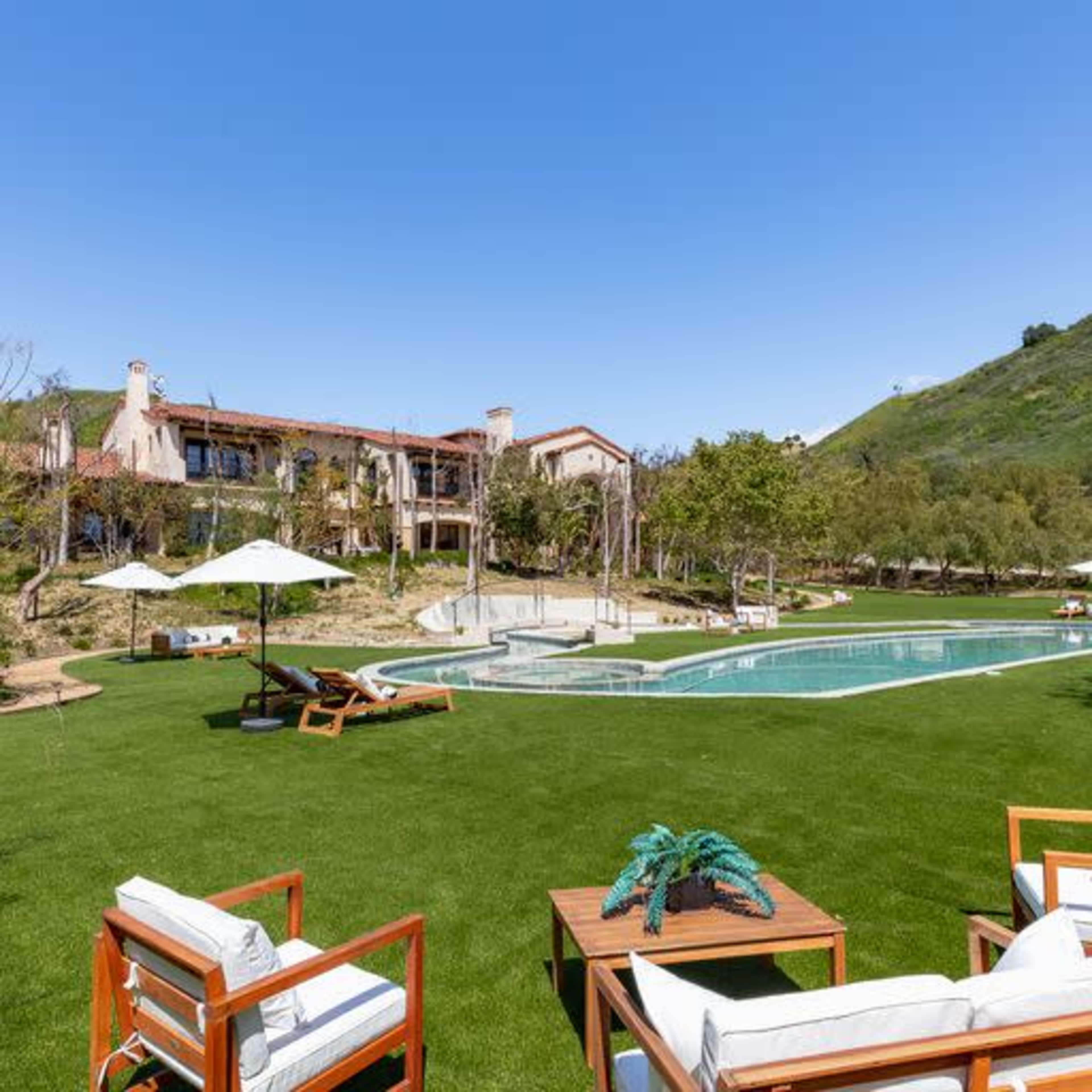 A spacious lawn area with lounge chairs and an umbrella near a large swimming pool, adjacent to a multi-level building surrounded by trees and hills.