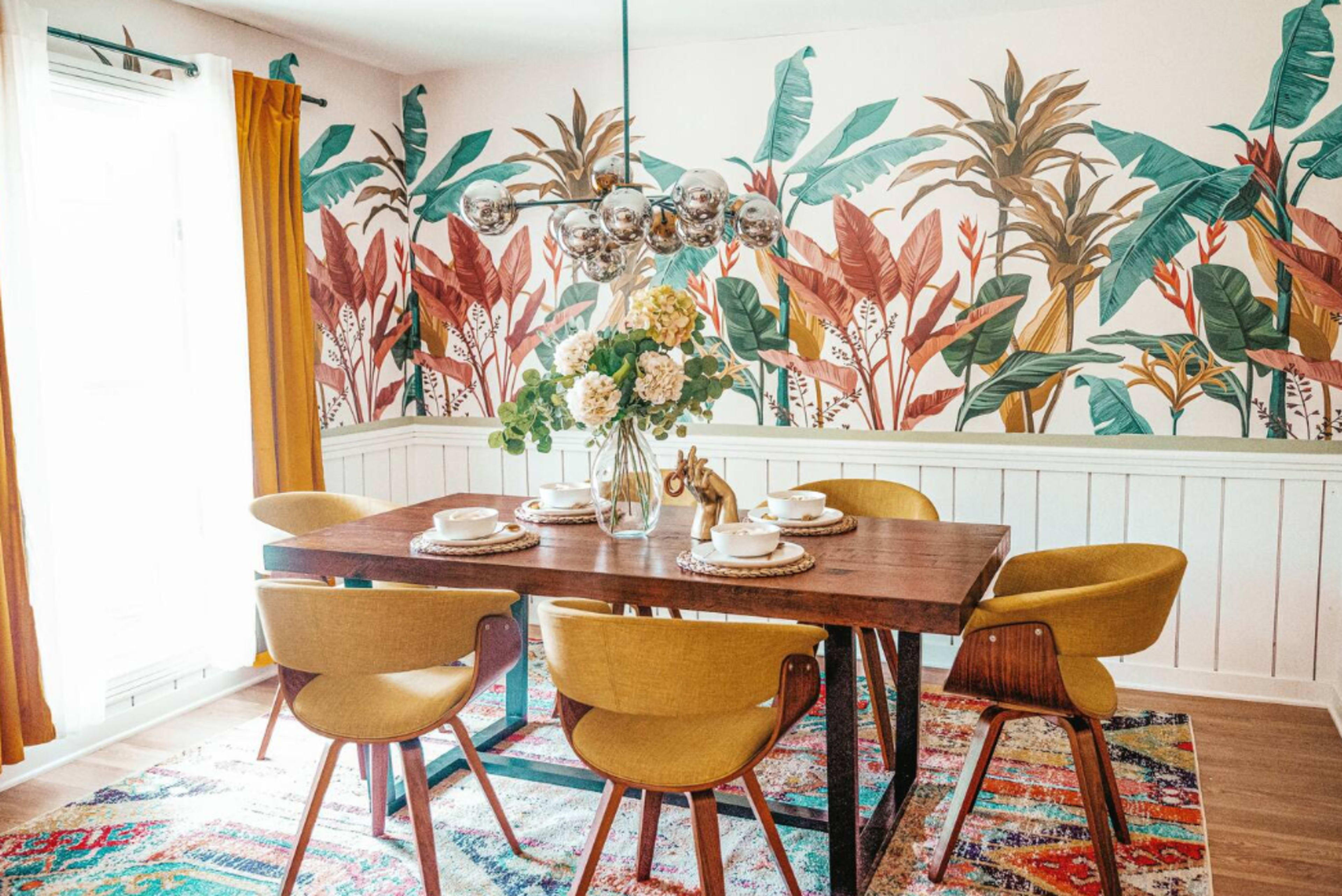 A dining room features a wooden table surrounded by yellow chairs, with a floral centerpiece and a colorful rug, set against a patterned wall of tropical plants.