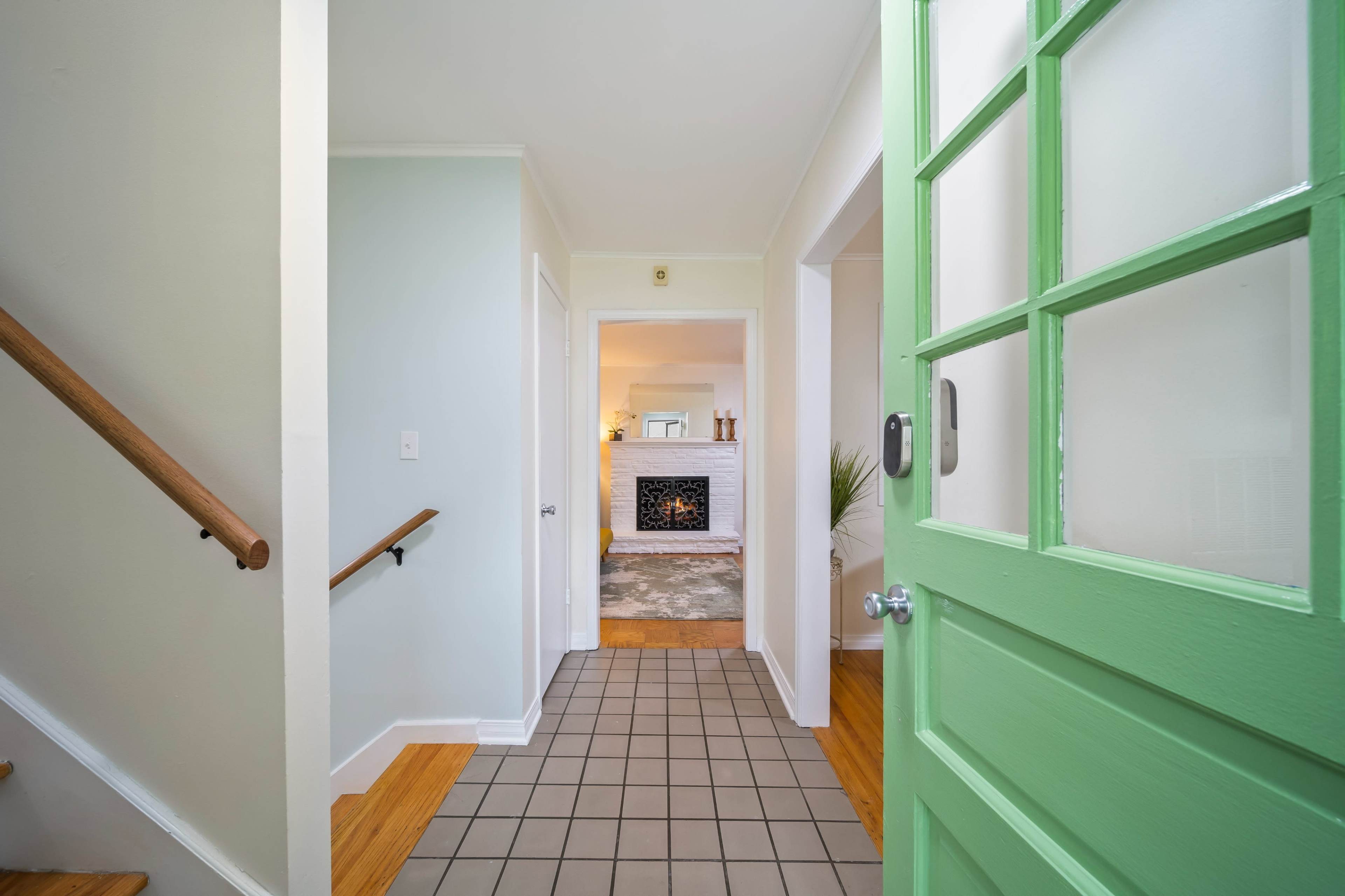A hallway with a green door, a staircase on the left, and an open view towards a living area featuring a fireplace.