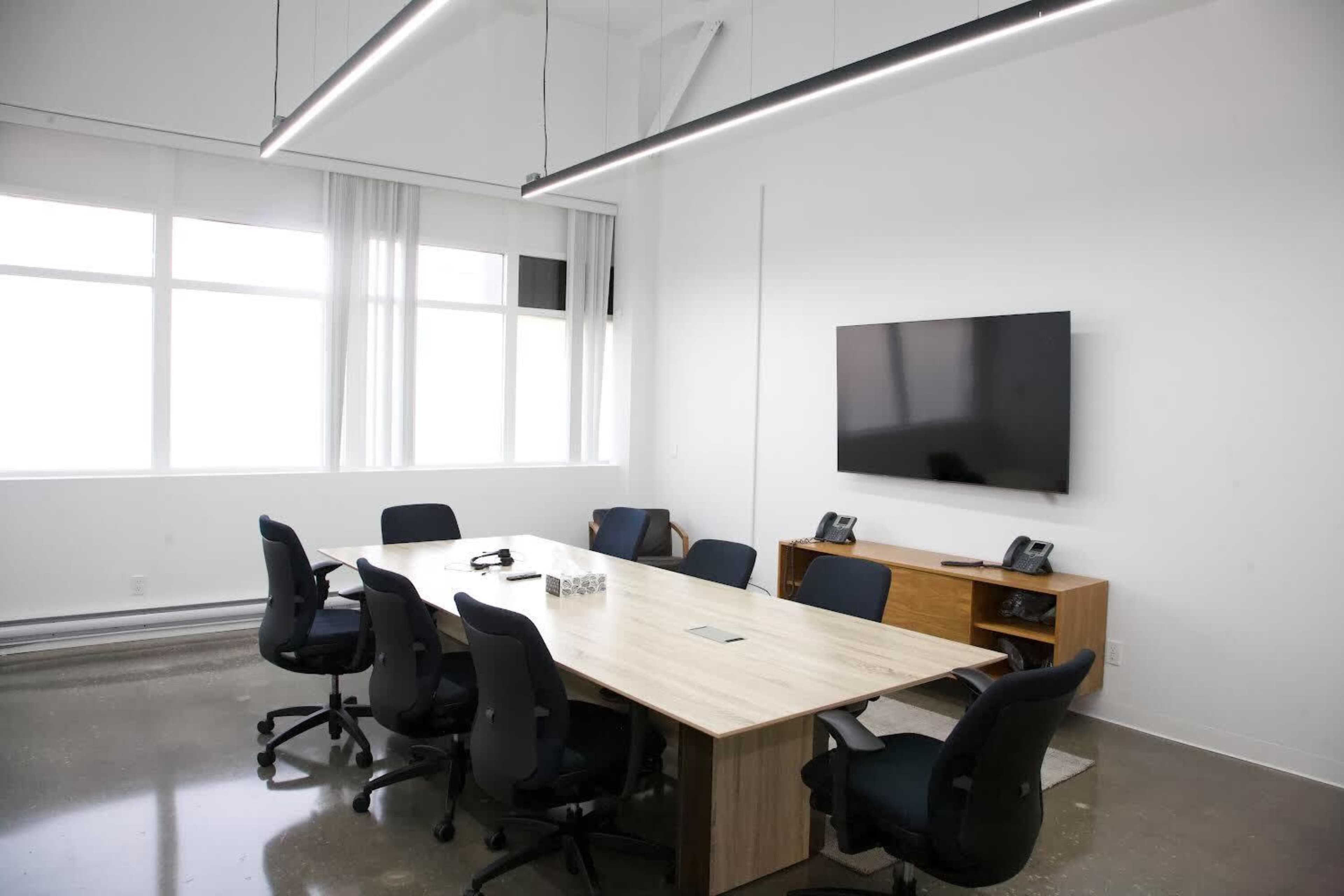 A modern conference room features a large wooden table surrounded by black chairs, with a wall-mounted television and minimal decor.