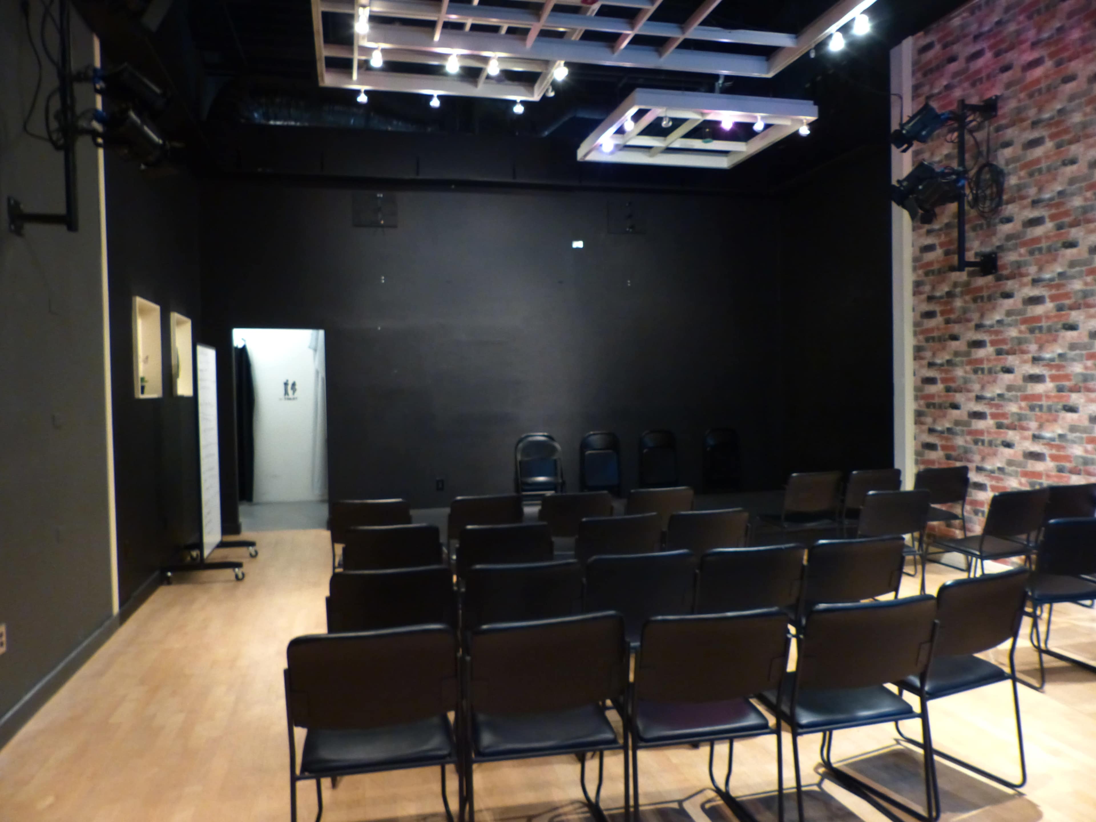 A small theater space with black walls, a row of black chairs facing a stage area, and a brick wall on one side.