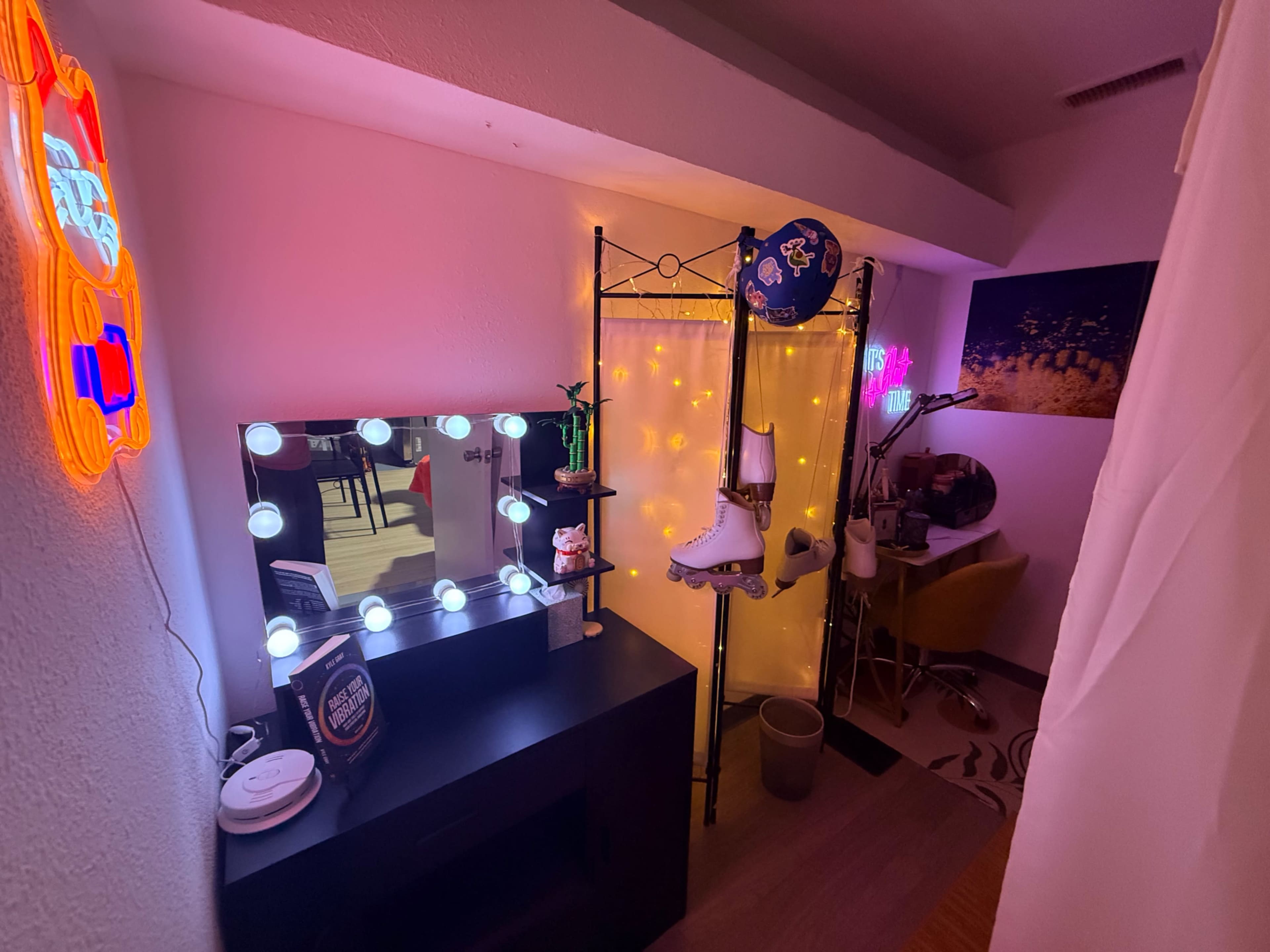 The image depicts a small room featuring a mirror with bright lights, decorative neon signage, a pair of roller skates hanging on a screen, and a desk with a chair.