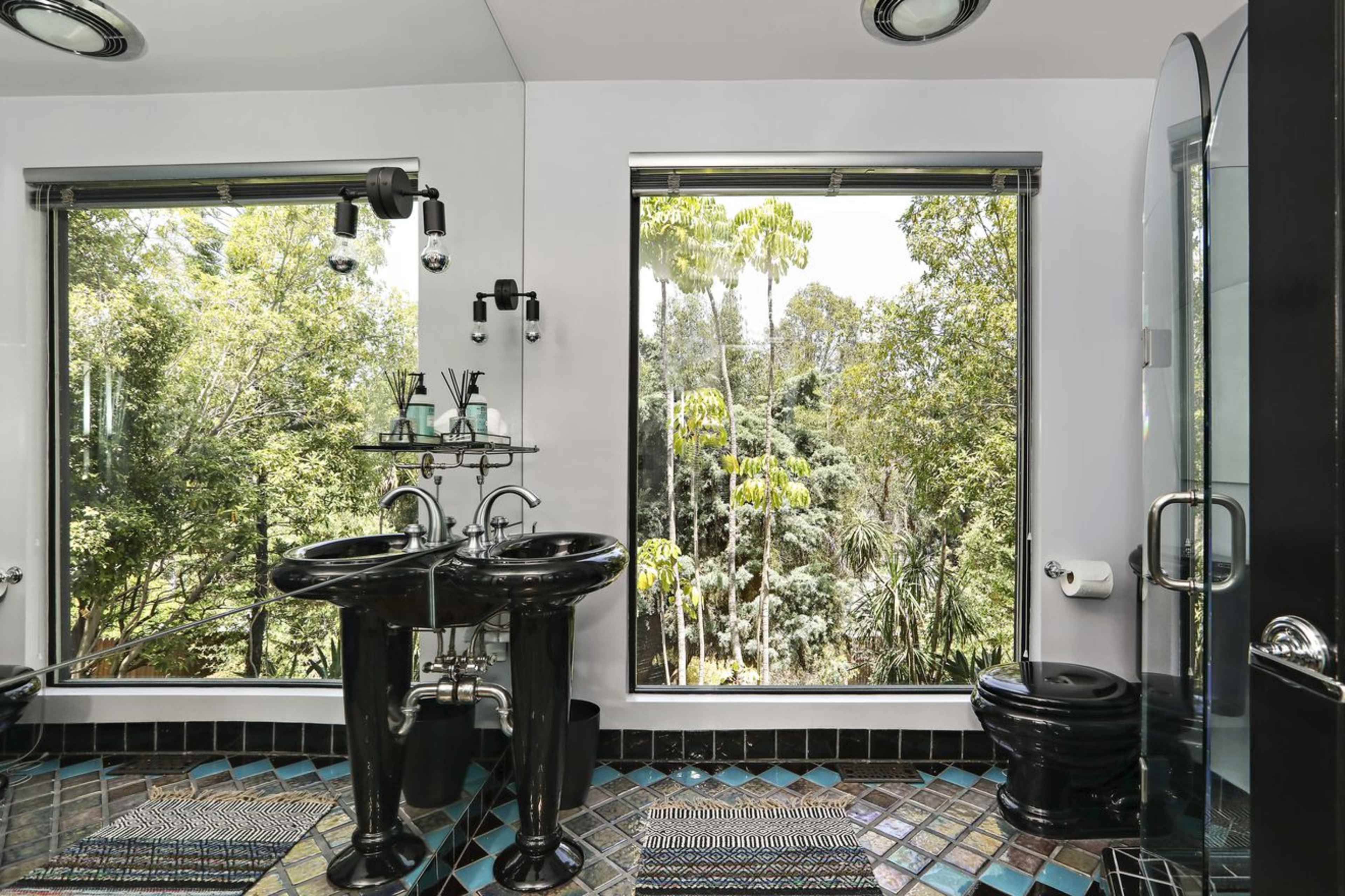 The bathroom features black sink basins and a toilet, with large windows overlooking a lush green landscape.