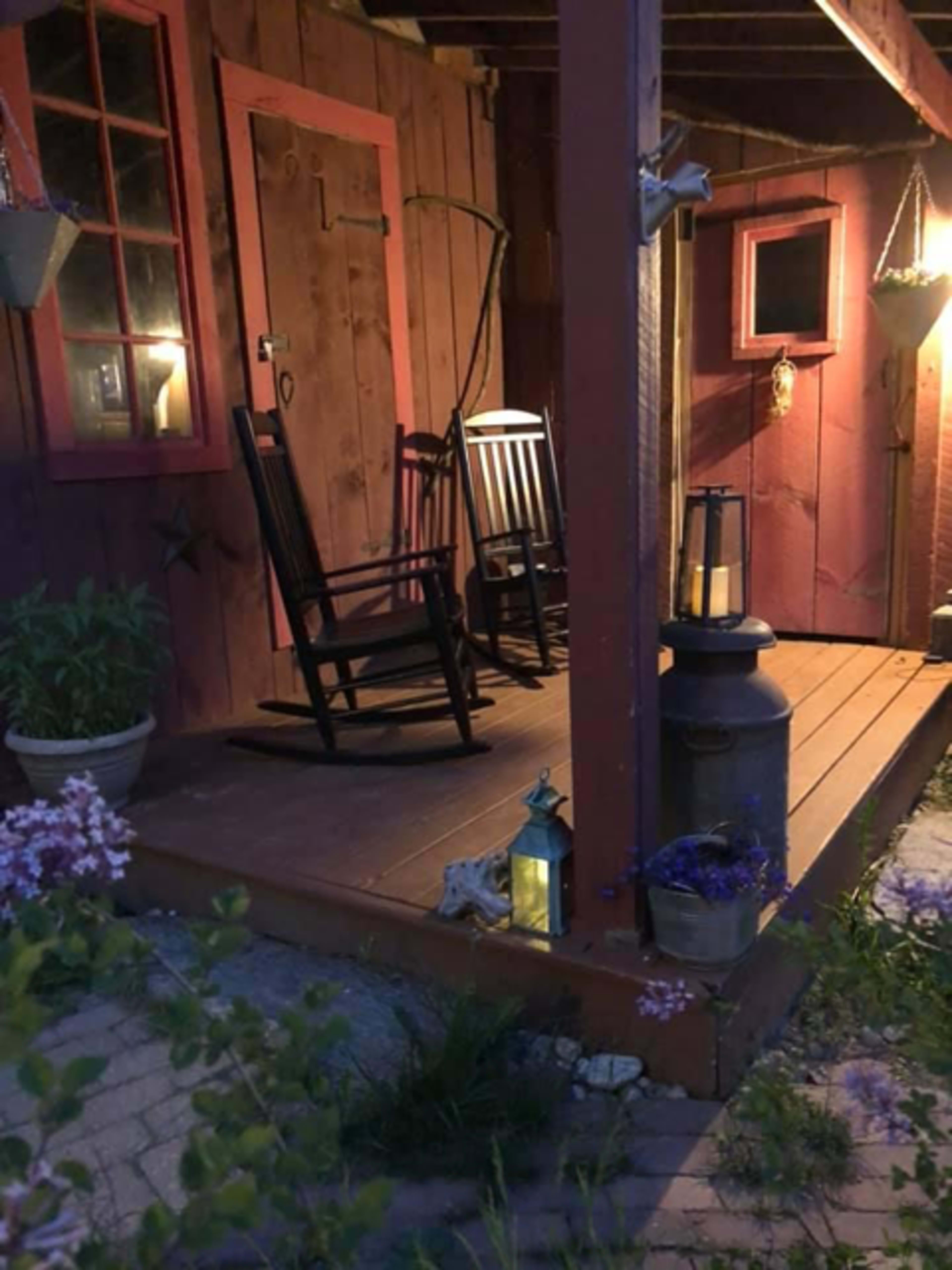 A rustic porch featuring two rocking chairs, potted plants, lanterns, and wooden walls with red trim.