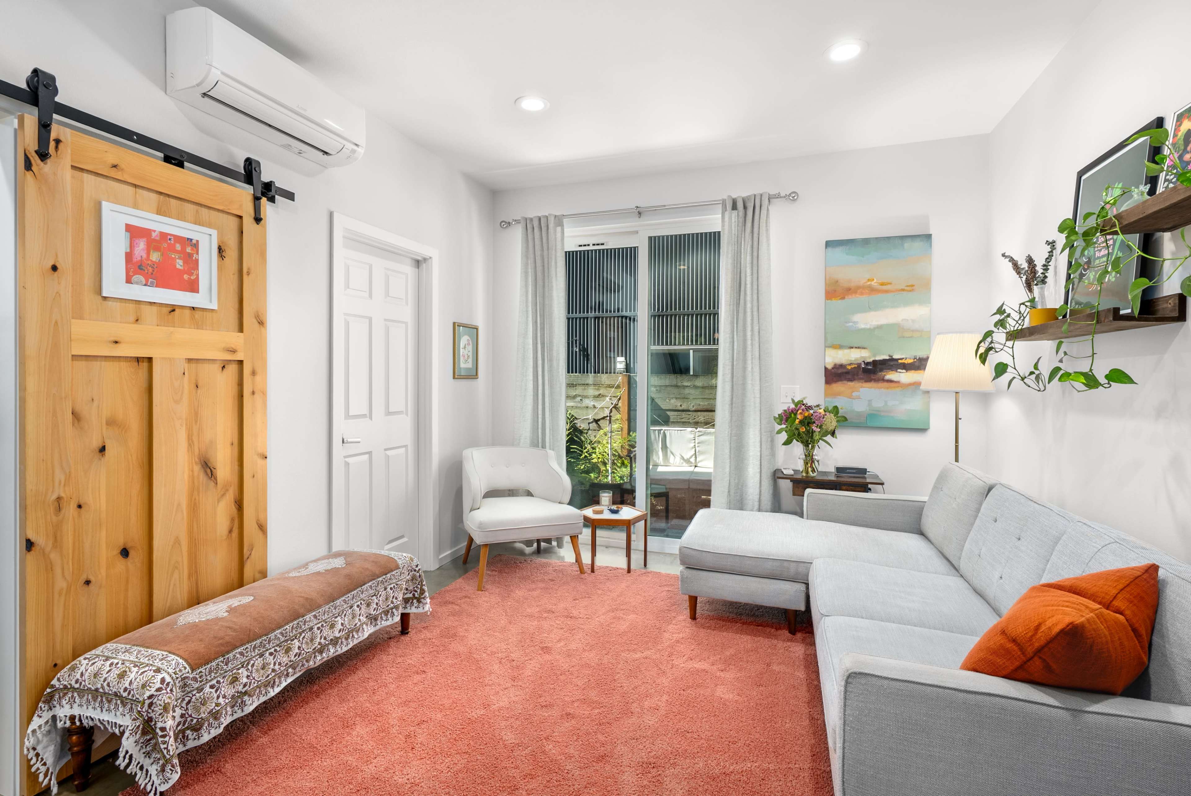 The image shows a cozy living room featuring a gray sectional sofa, a white armchair, a wooden sliding door, and large windows with light filtering through.