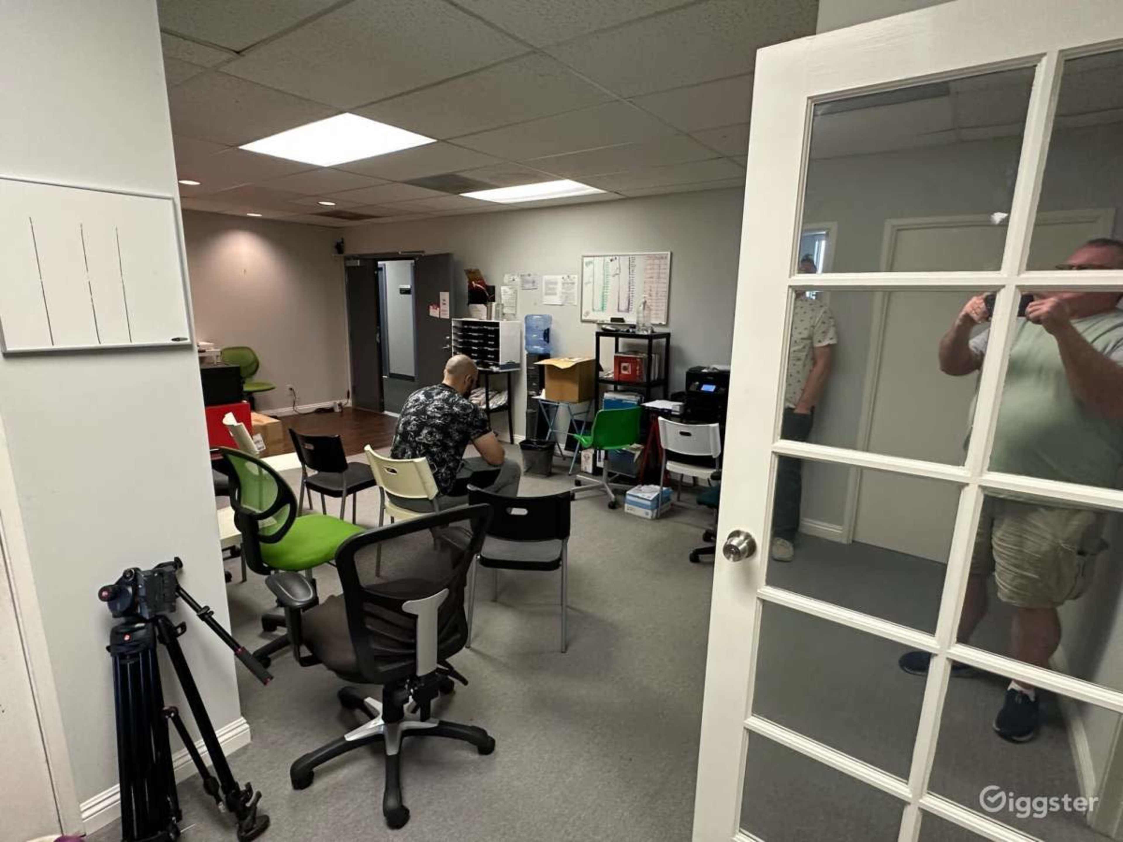 A small office space is shown with several chairs arranged around a central table, and a person sitting at a desk in the corner.
