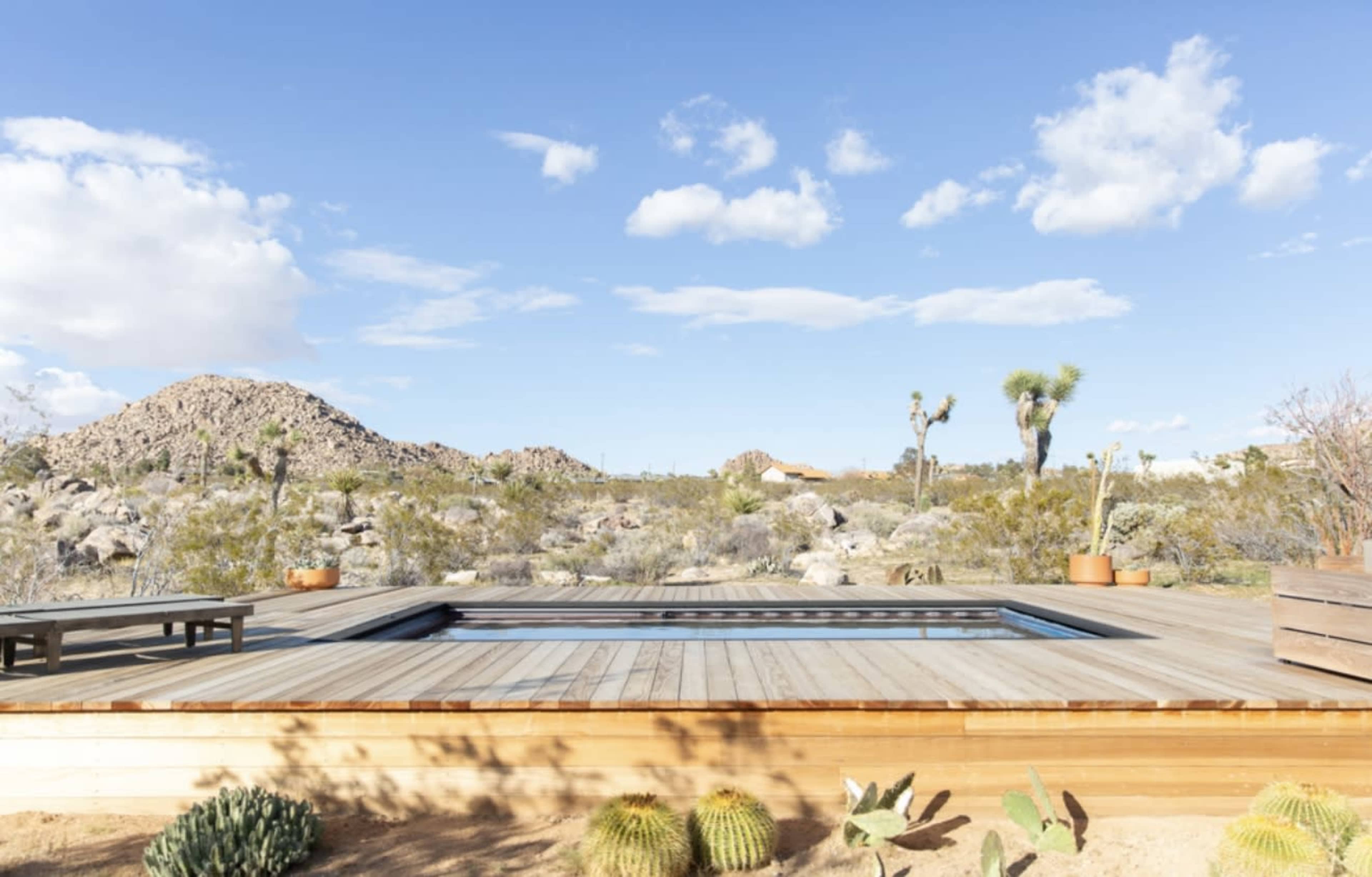The image shows a wooden deck with a rectangular pool surrounded by desert vegetation and rocky hills under a blue sky with scattered clouds.