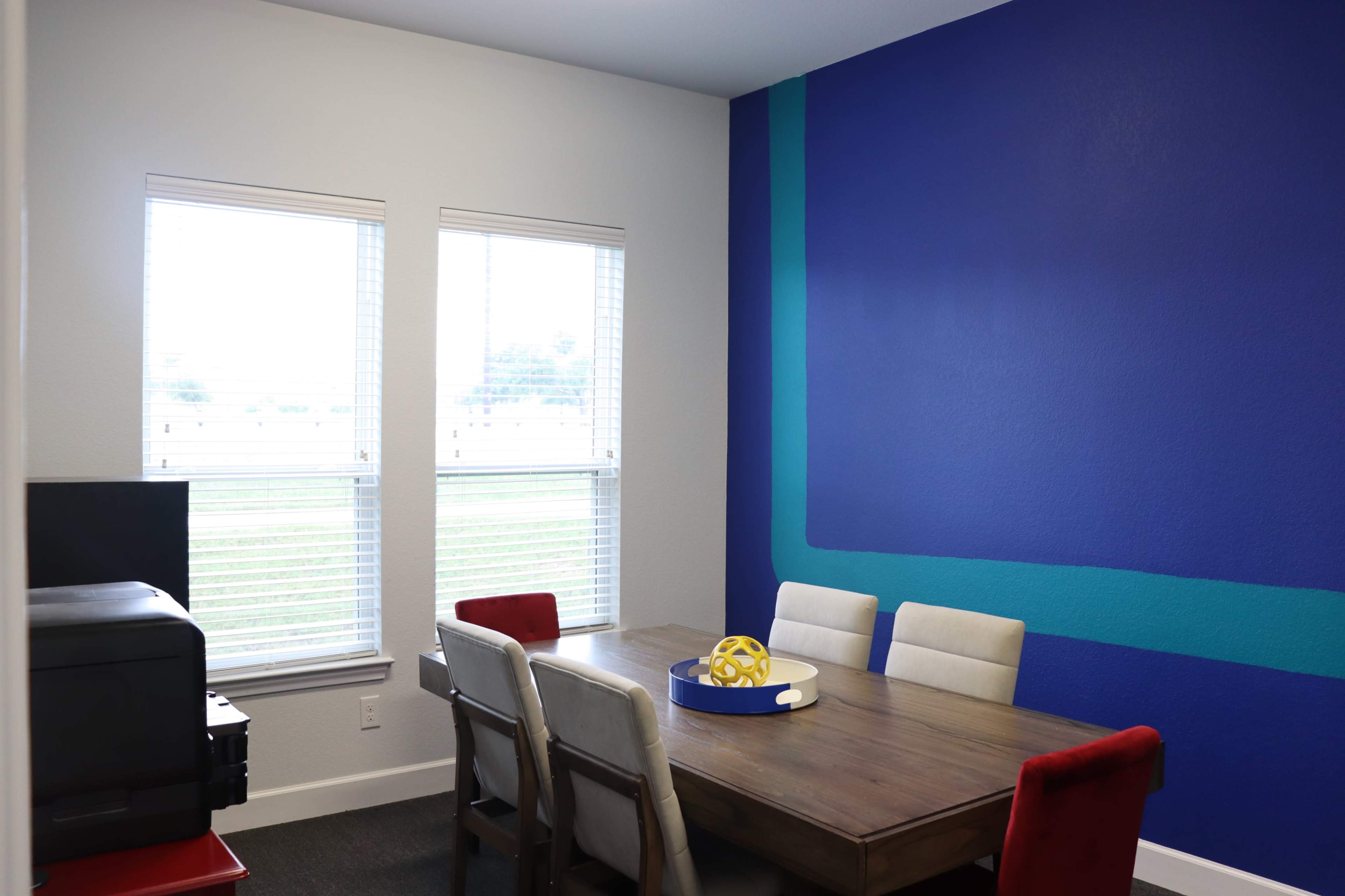 A dining area features a wooden table surrounded by six chairs, with large windows allowing natural light and a blue accent wall.