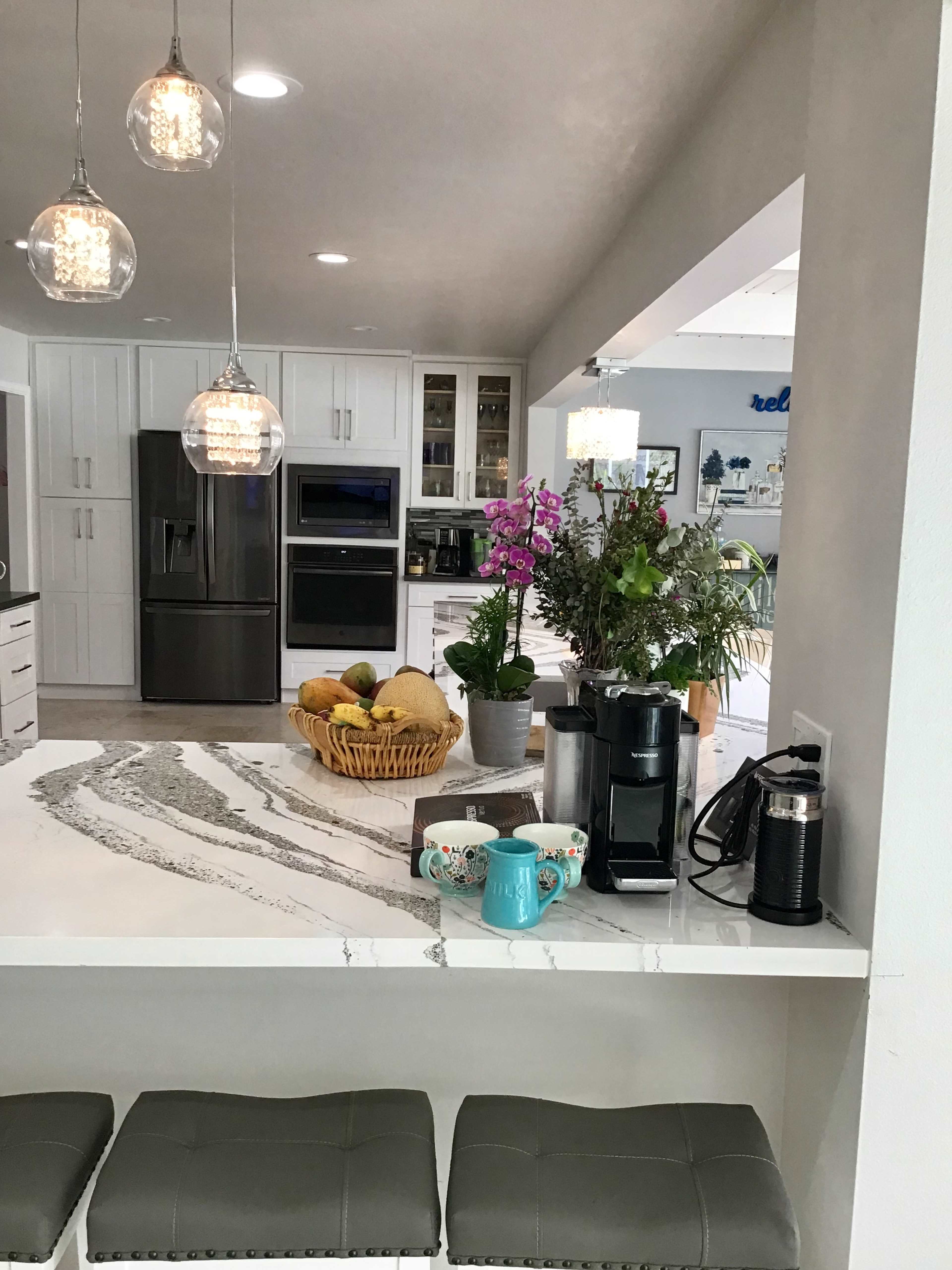 A modern kitchen features a marble countertop with a coffee maker and colorful mugs, surrounded by pendant lighting and various plants.