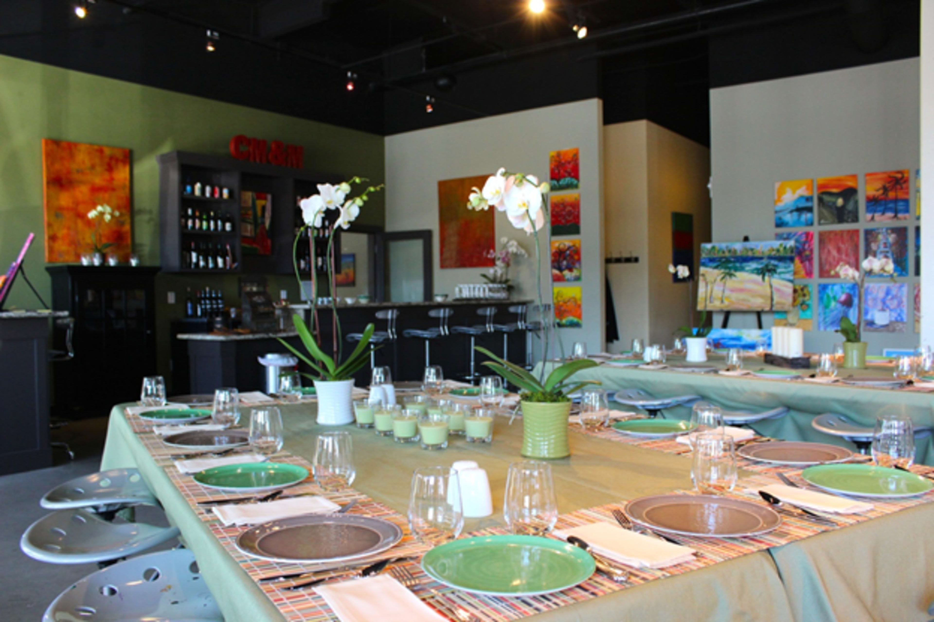 A modern restaurant interior with a long dining table set with green plates, glasses, and decorative plants, against a backdrop of colorful artwork.