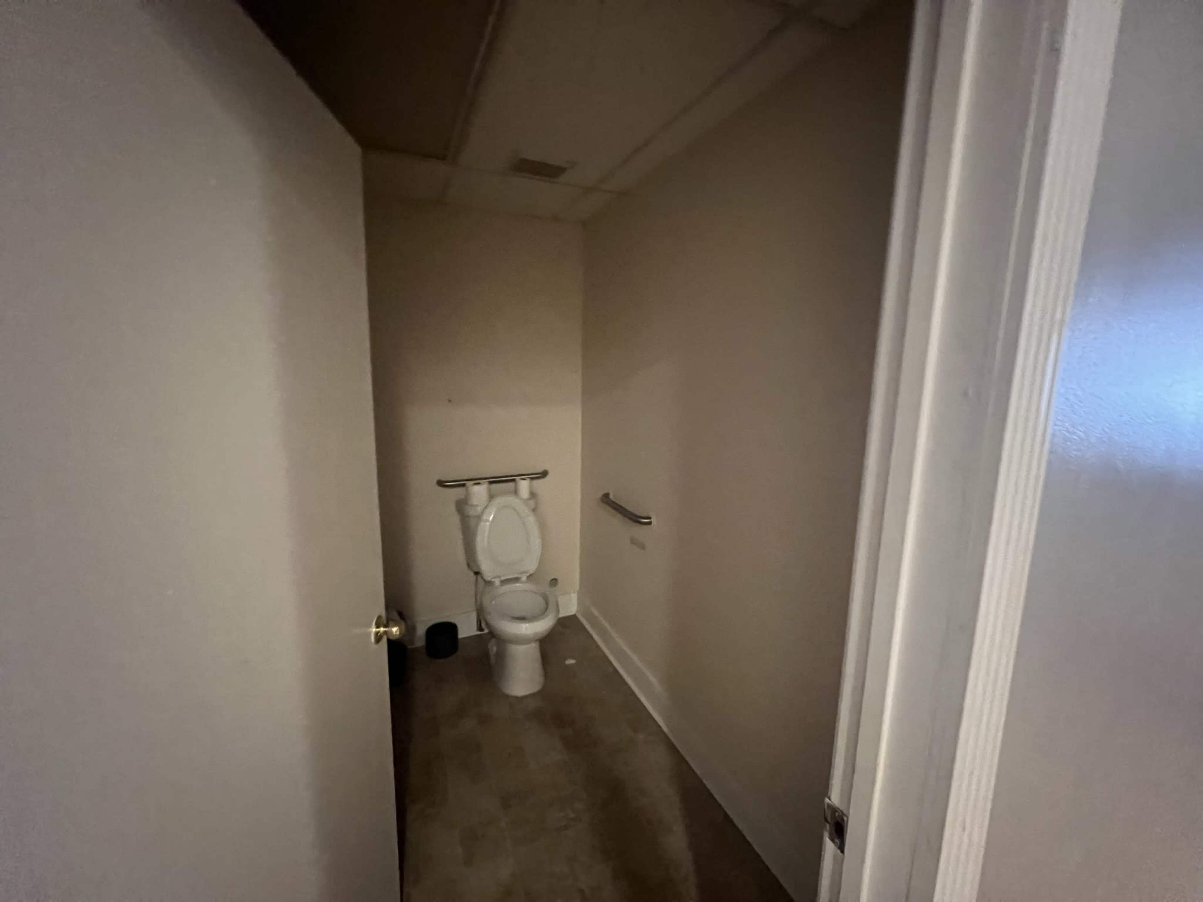 A small, dimly lit bathroom features a toilet and a handrail against a plain wall.