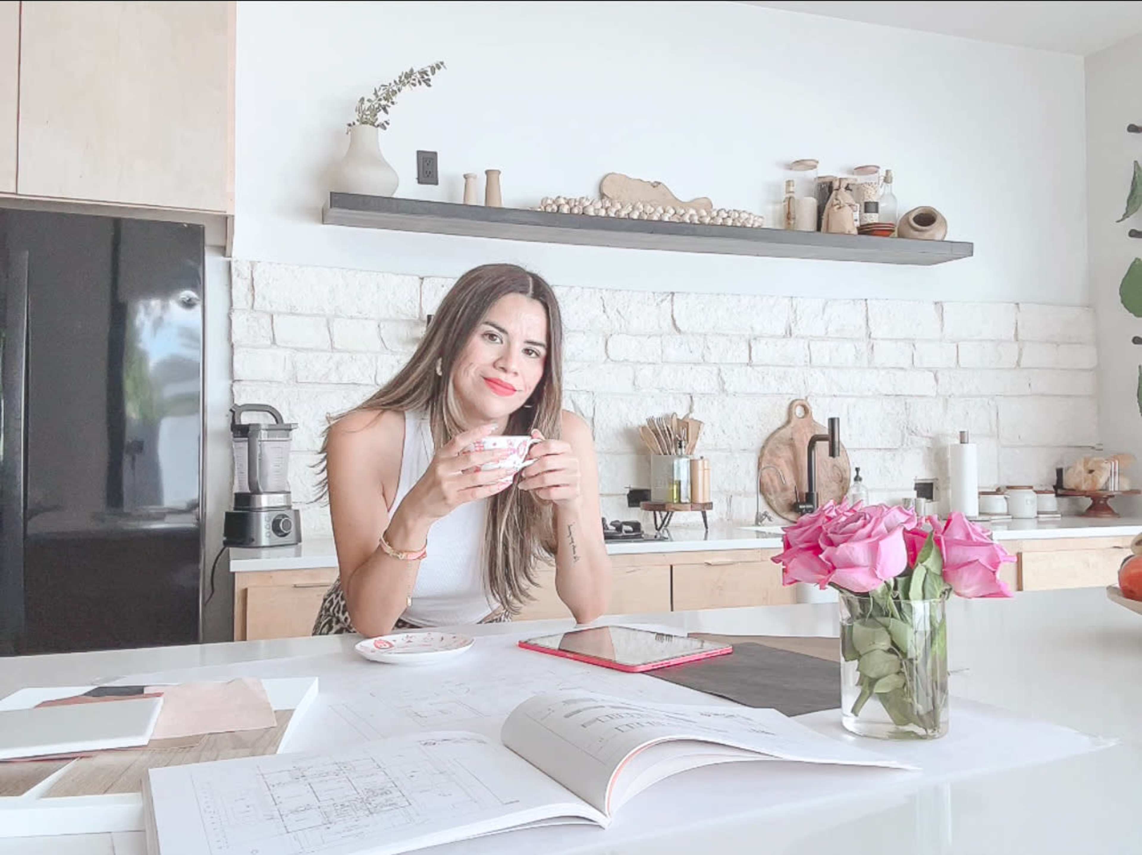 A woman sits at a modern kitchen table, holding a cup, with pink roses in a vase and various kitchen items displayed on a shelf behind her.