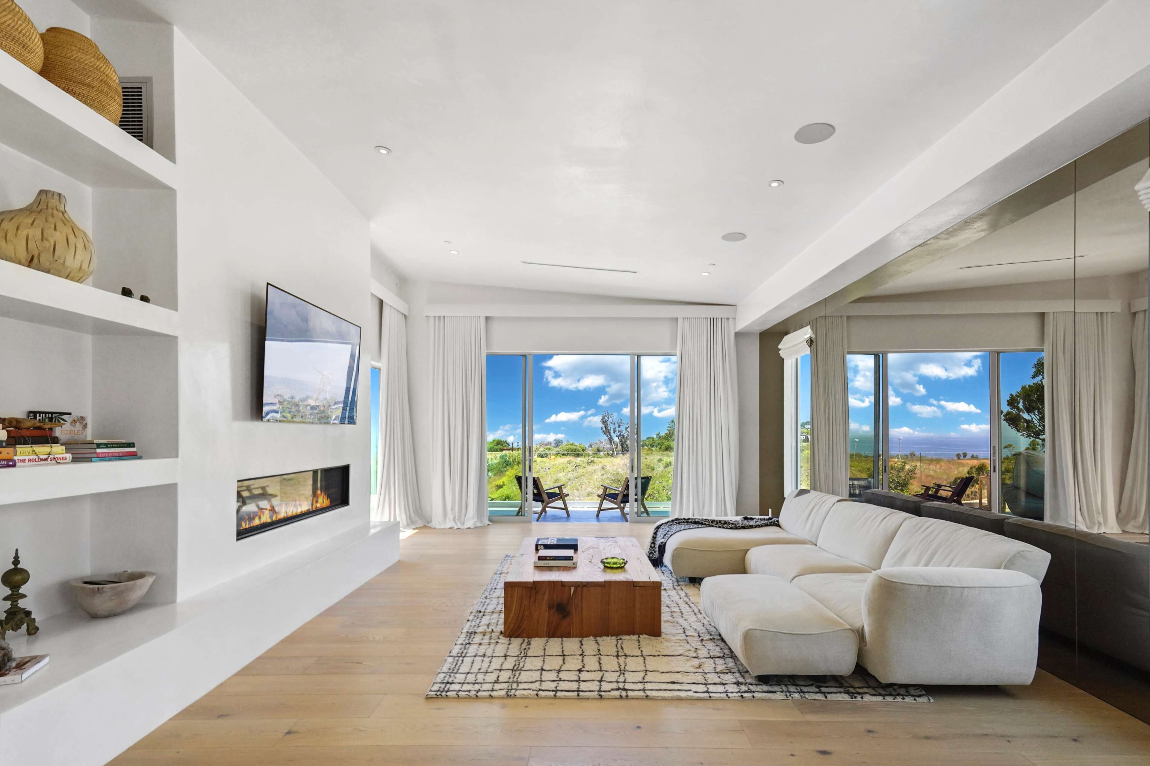 The living room features a white sectional sofa, a wooden coffee table, and large windows with views of greenery and blue sky.