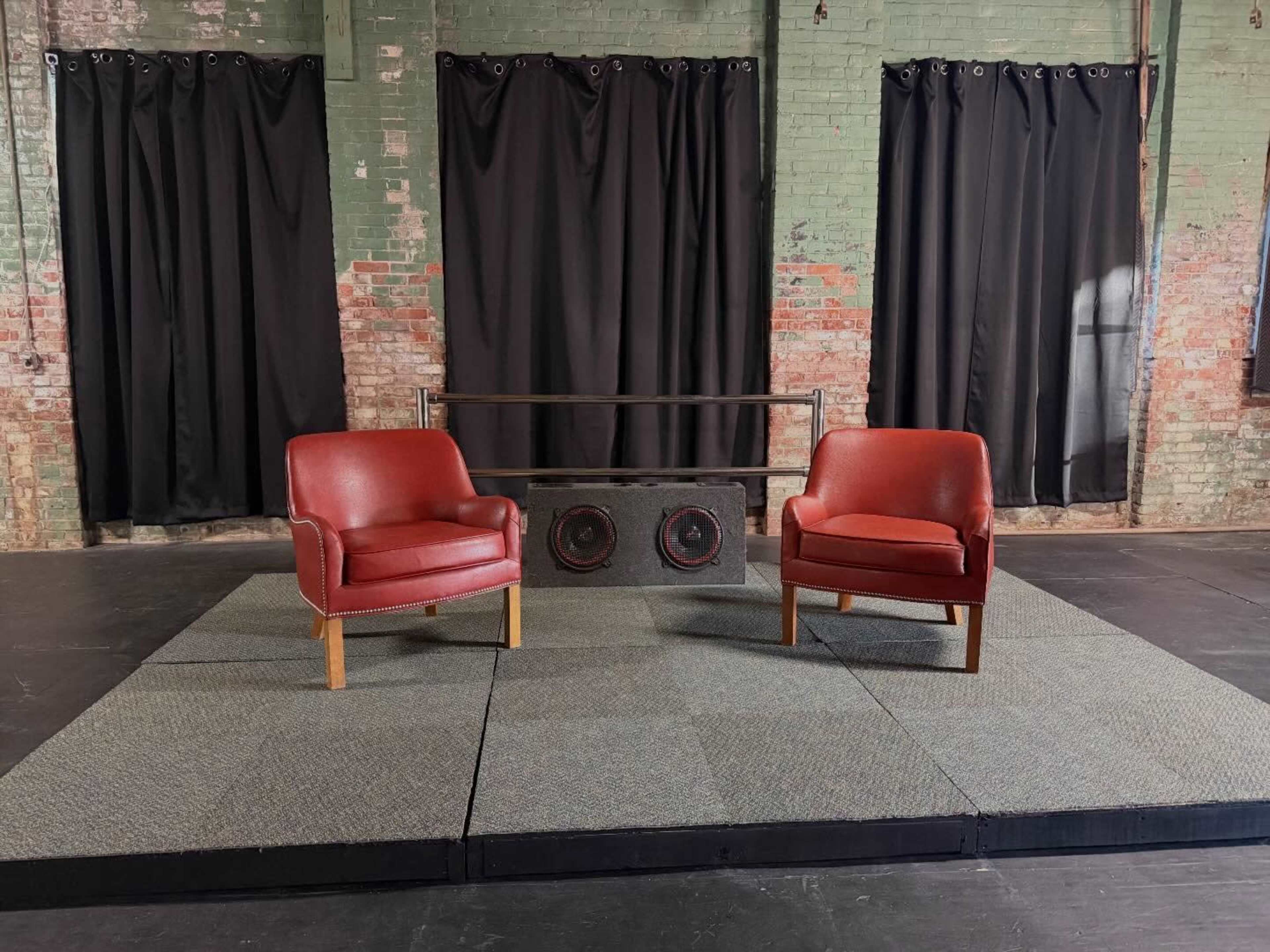 The image shows a stage with two red chairs placed on a carpeted platform, flanked by black curtains against a brick wall.