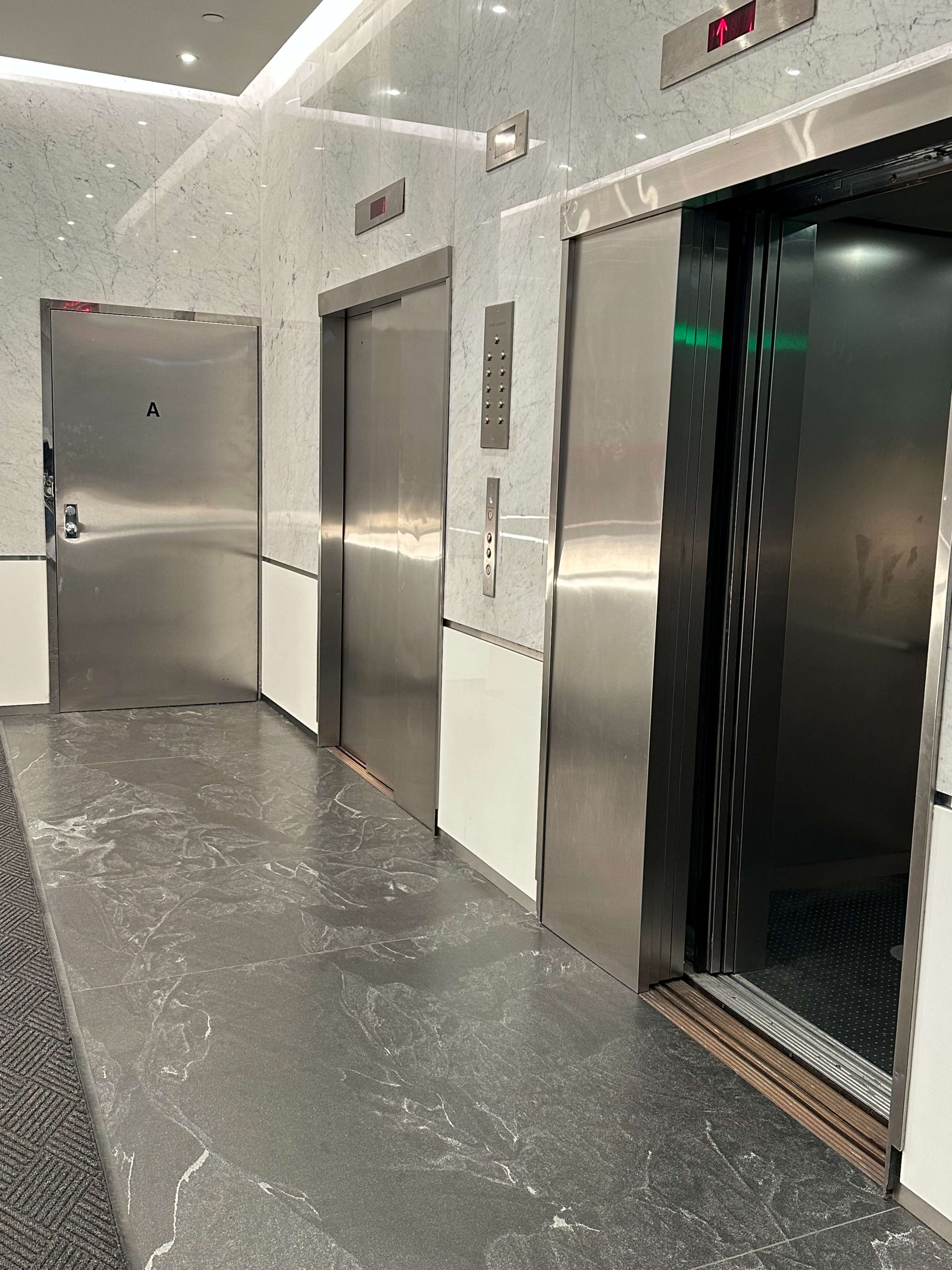 The image shows an elevator lobby with two elevators and a door, featuring a marble wall and a textured floor.