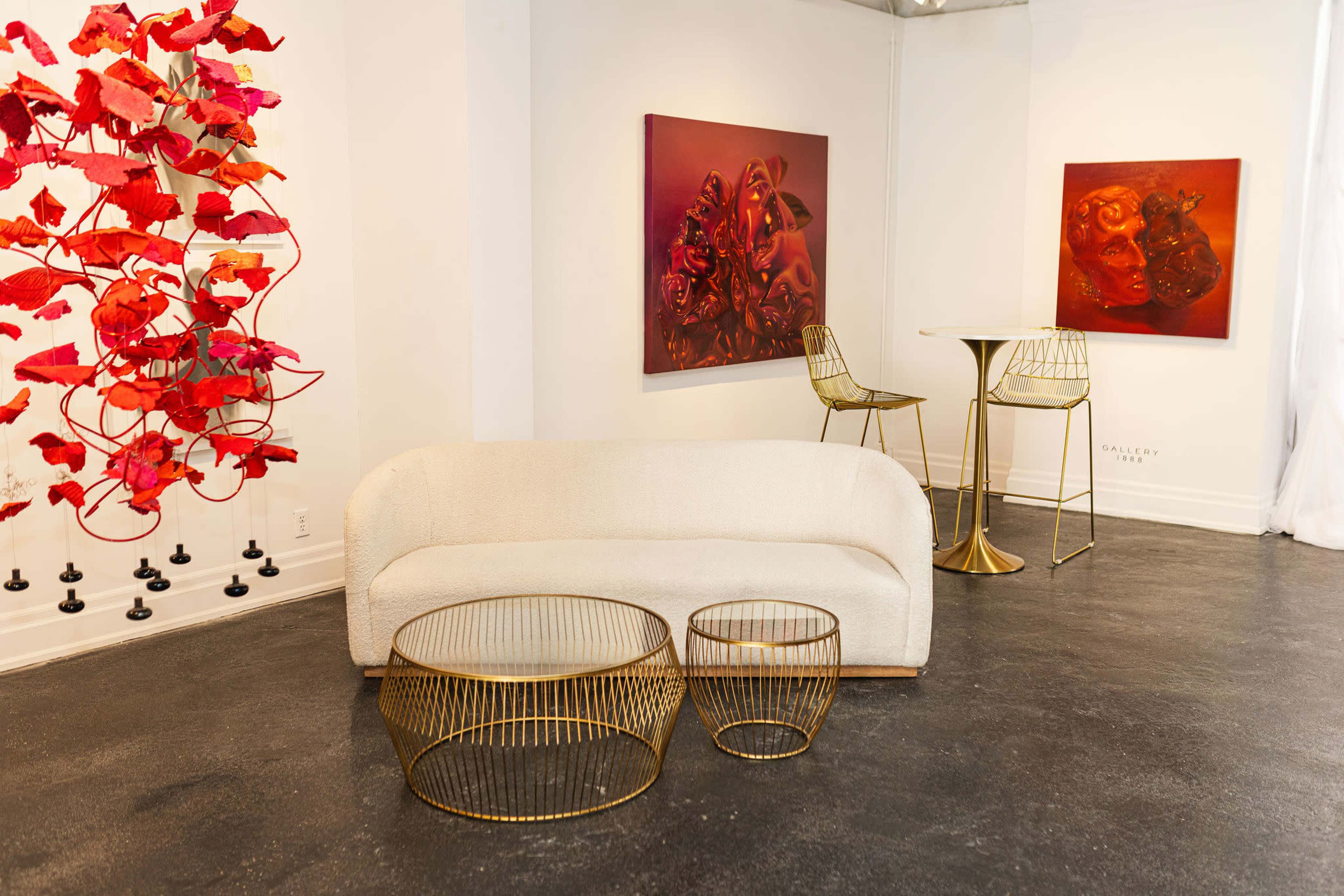 The image shows a modern art gallery featuring a white sofa, metallic tables, and red-themed artworks on the walls.