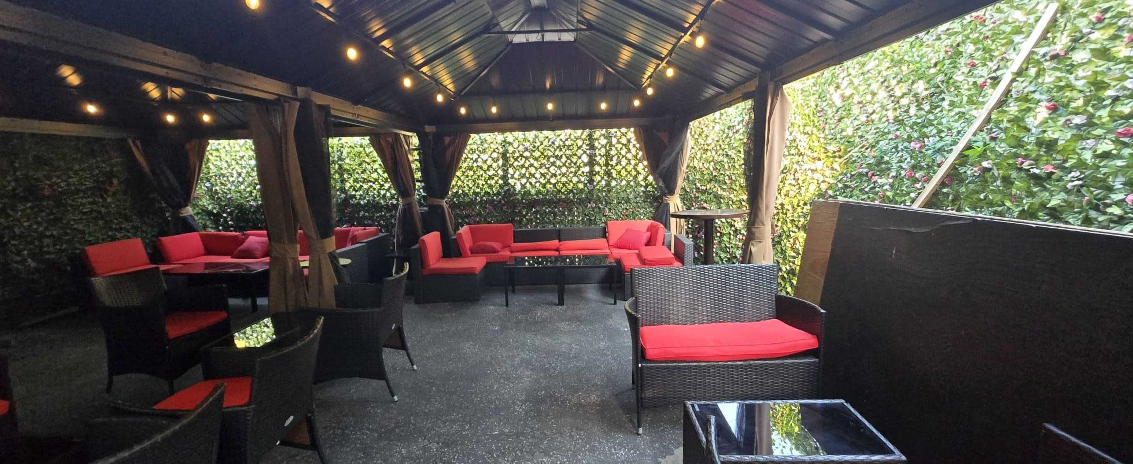 The image shows a covered outdoor seating area with red cushioned furniture and greenery surrounding the space.