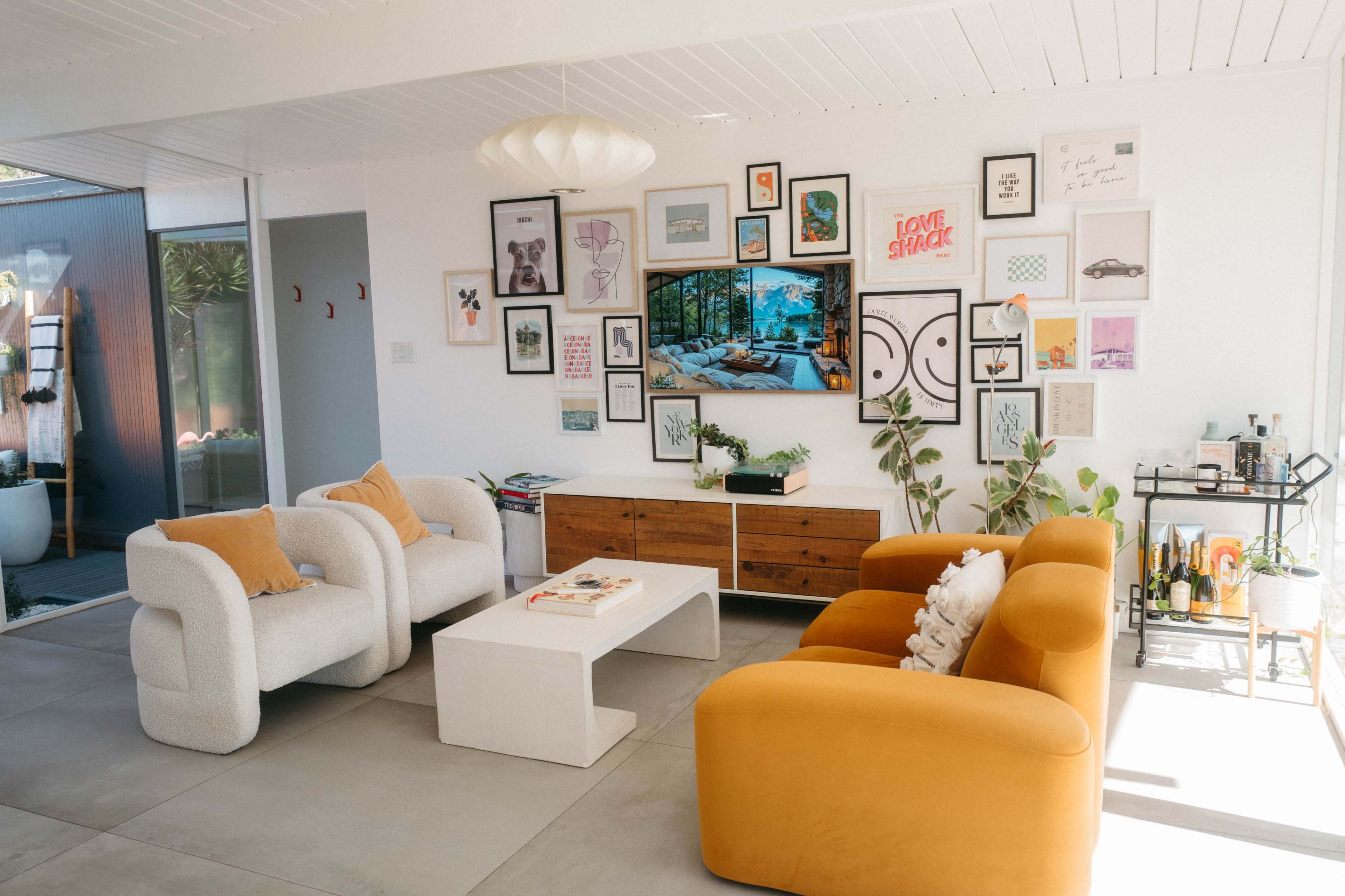 A modern living room features a white wall adorned with a gallery of framed artwork, while two textured armchairs and a yellow sofa surround a minimalist coffee table.