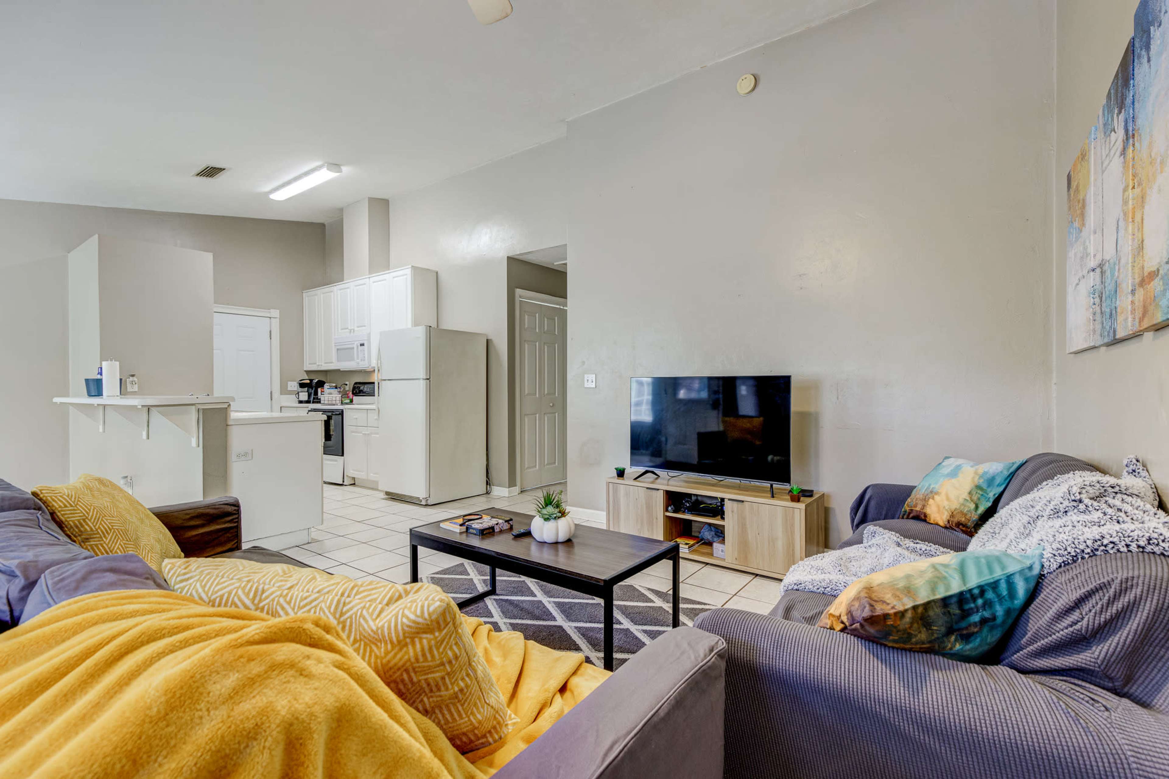 A living room features two sofas, a coffee table, and a television, with a kitchen visible in the background.