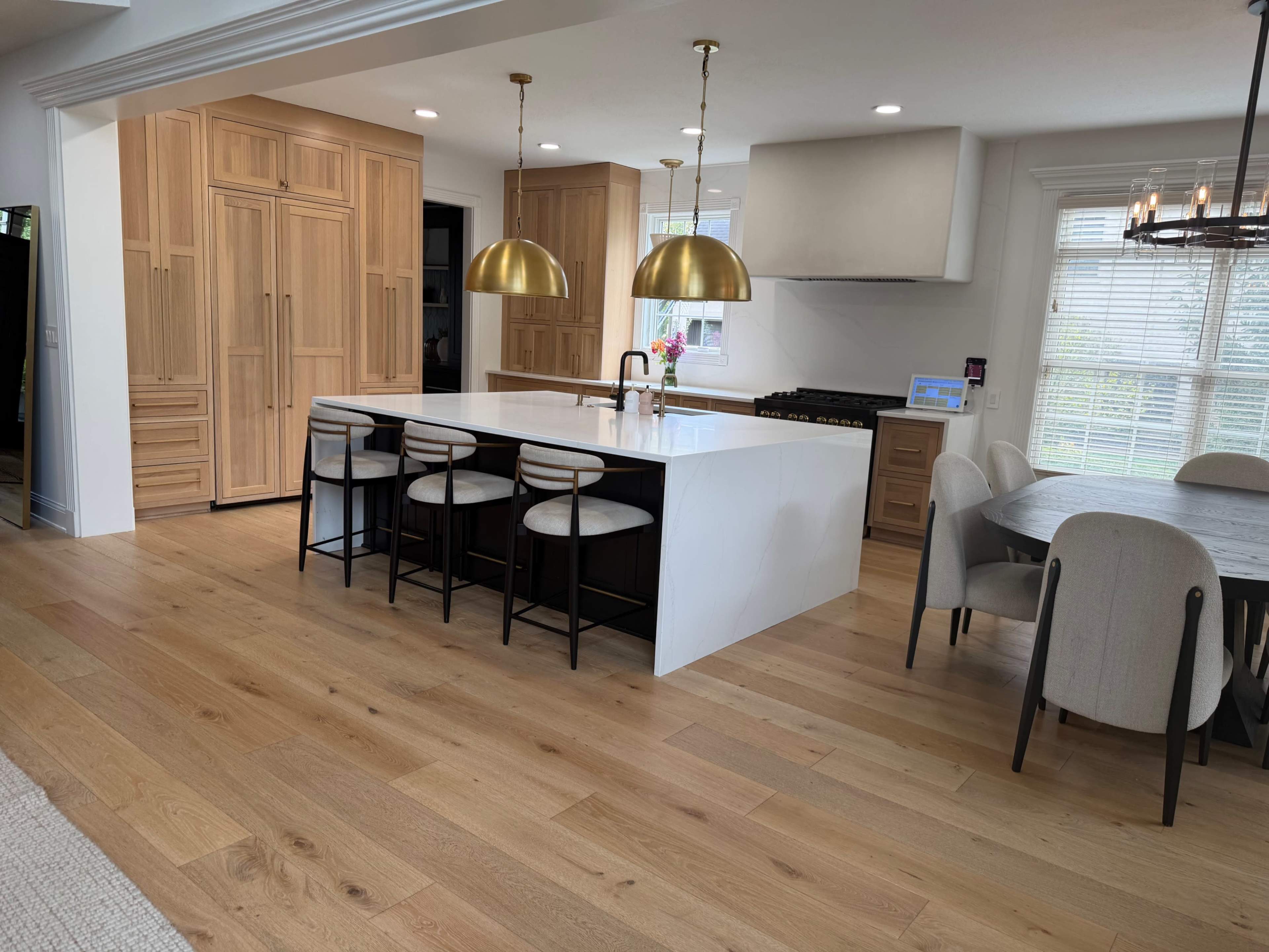The image shows a modern kitchen featuring light wood cabinetry, a large white island with seating for four, and two gold pendant lights overhead.