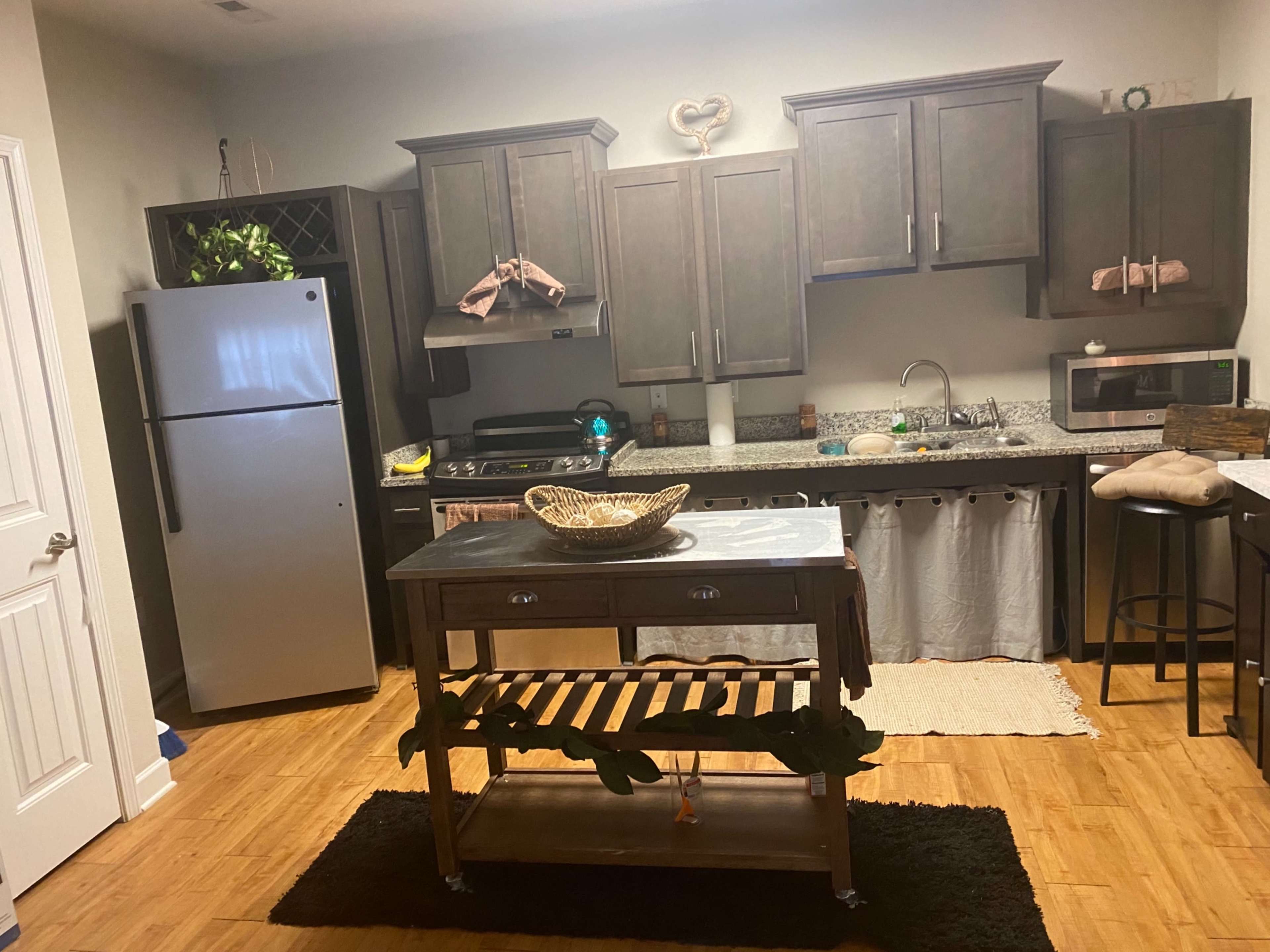A kitchen with dark cabinets, a silver refrigerator, a wooden island with a decorative bowl, and a black rug on a wood floor.