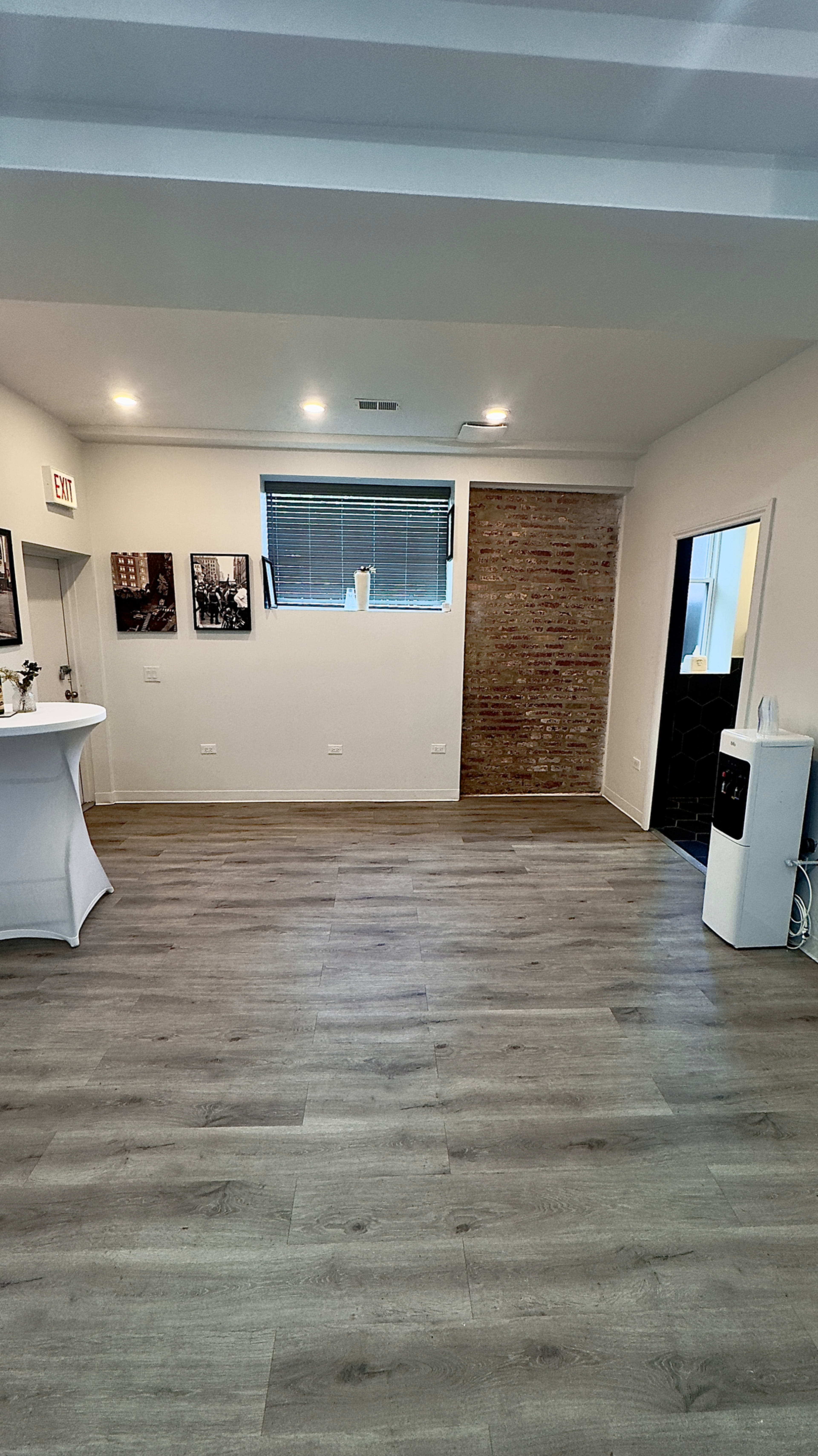 The image shows a spacious room with wooden flooring, a few framed pictures on the walls, and a small white table in one corner.