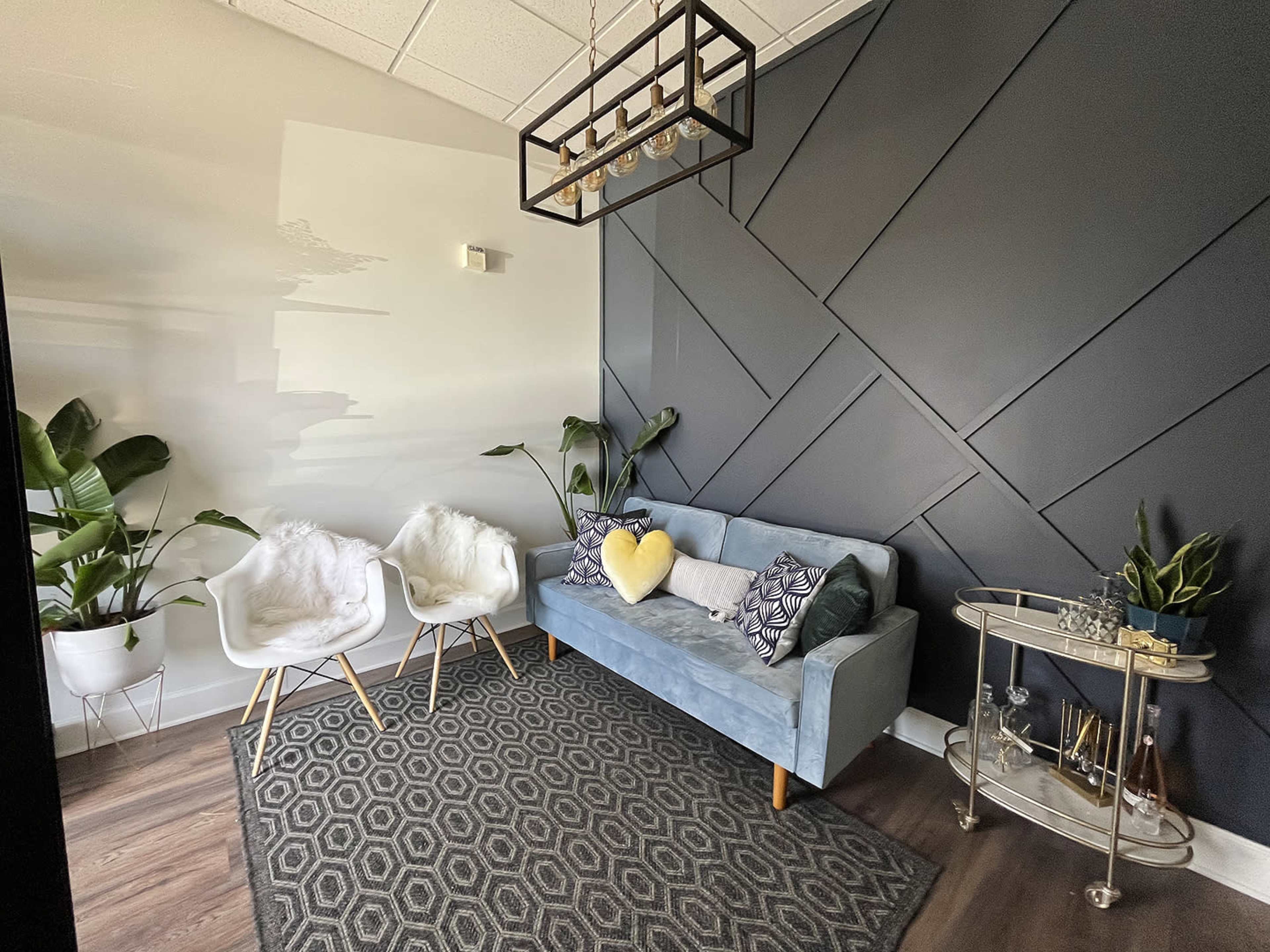 A small, modern seating area featuring a blue sofa, two white chairs, and a geometric-patterned rug, with plants and a glass table in the background.