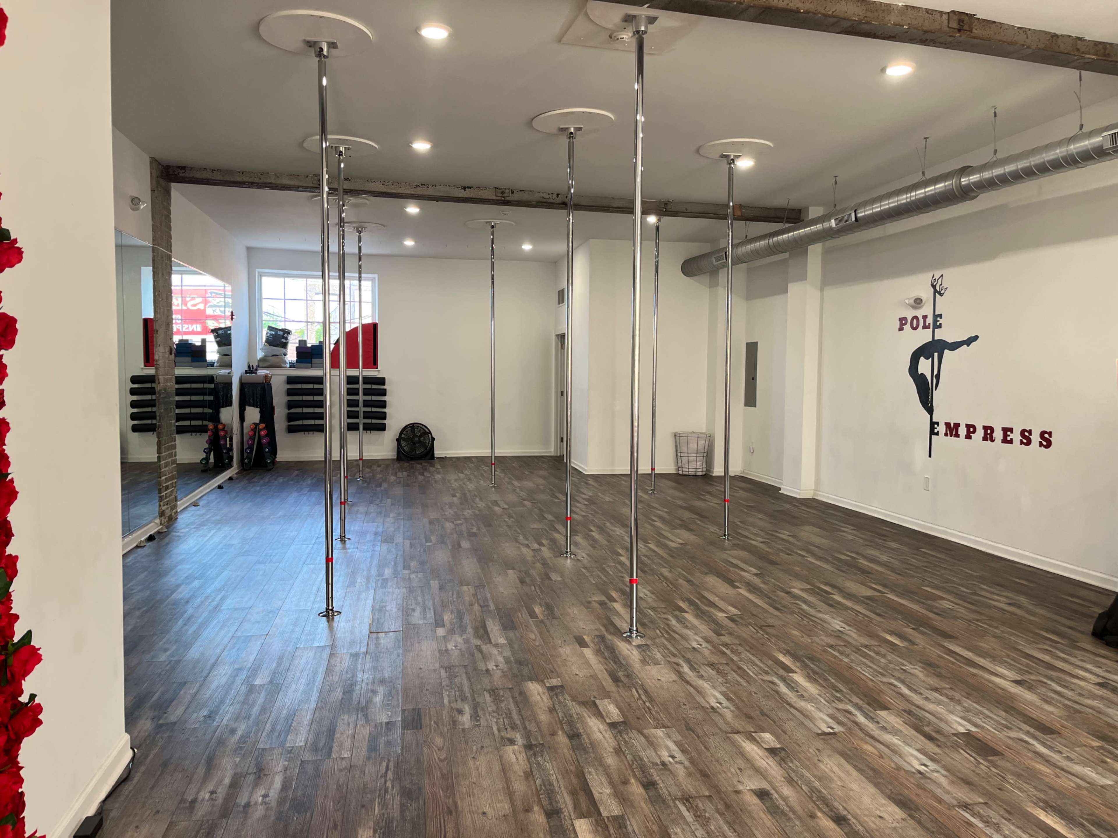 The image shows a spacious dance studio with several vertical poles and a wooden floor, featuring white walls and a small fan in one corner.