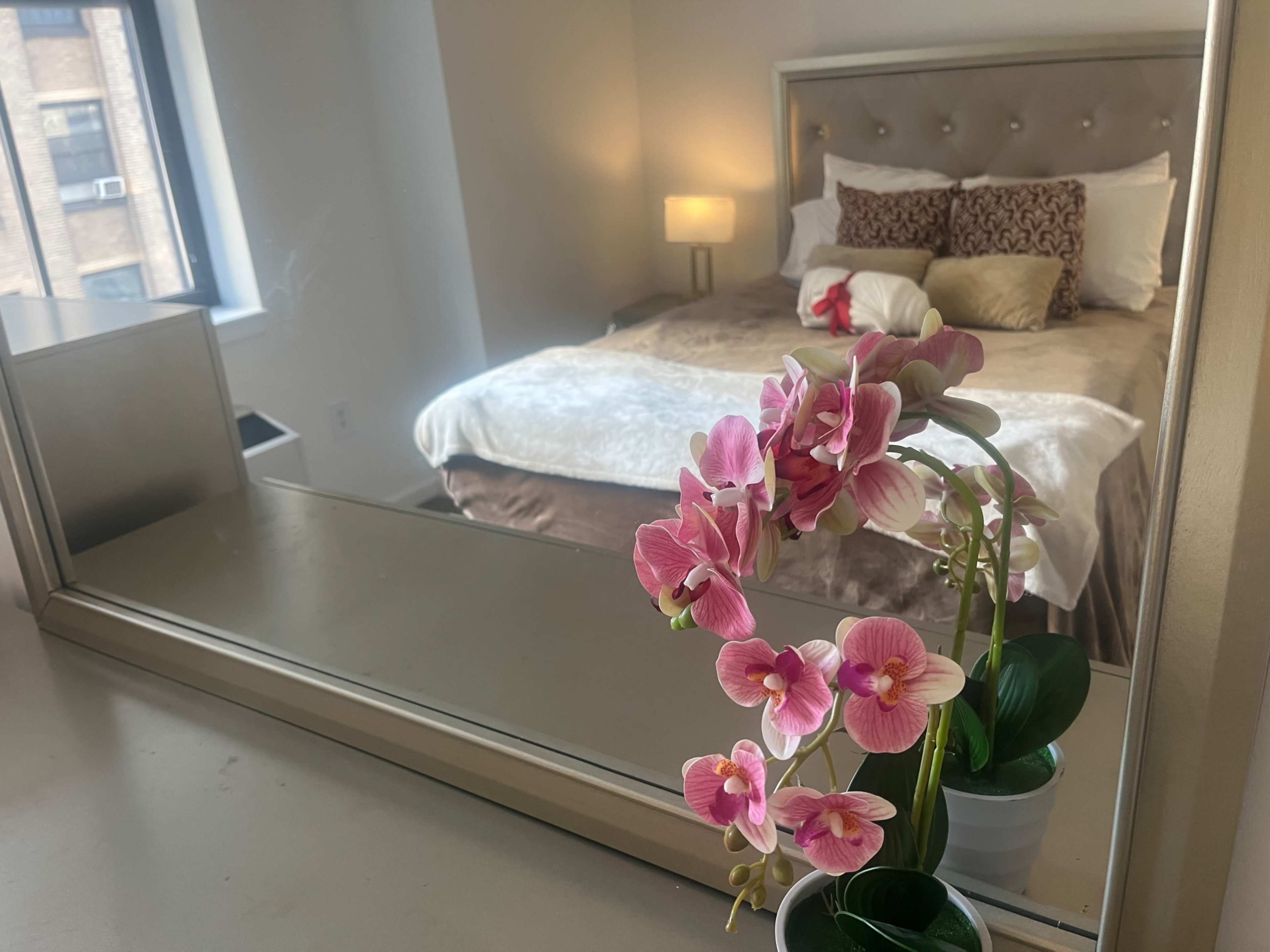 A bedroom features a neatly made bed with decorative pillows and a bedside lamp, while a potted orchid is reflected in a mirror on the dresser.