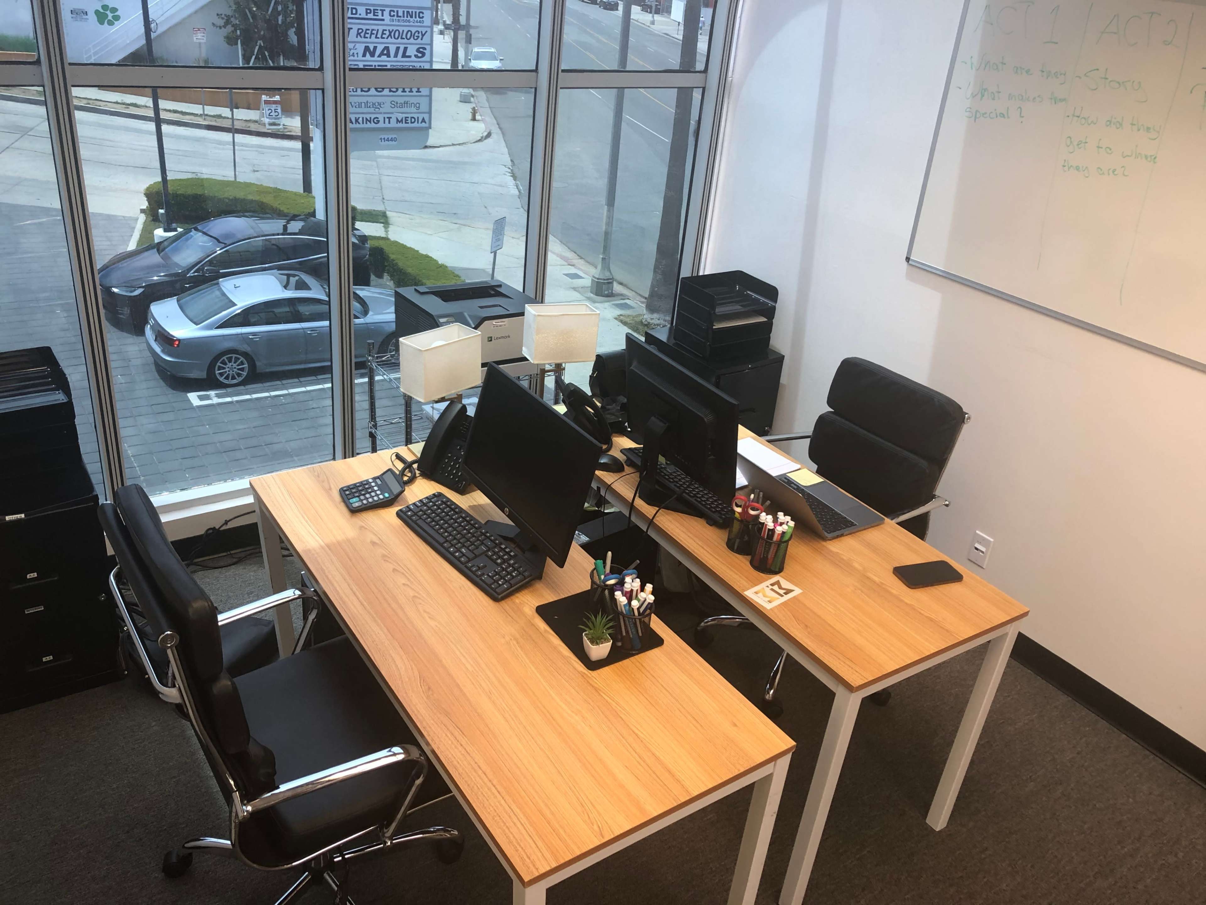A modern office space with two wooden desks, each equipped with computers and office supplies, located near large windows overlooking a street.
