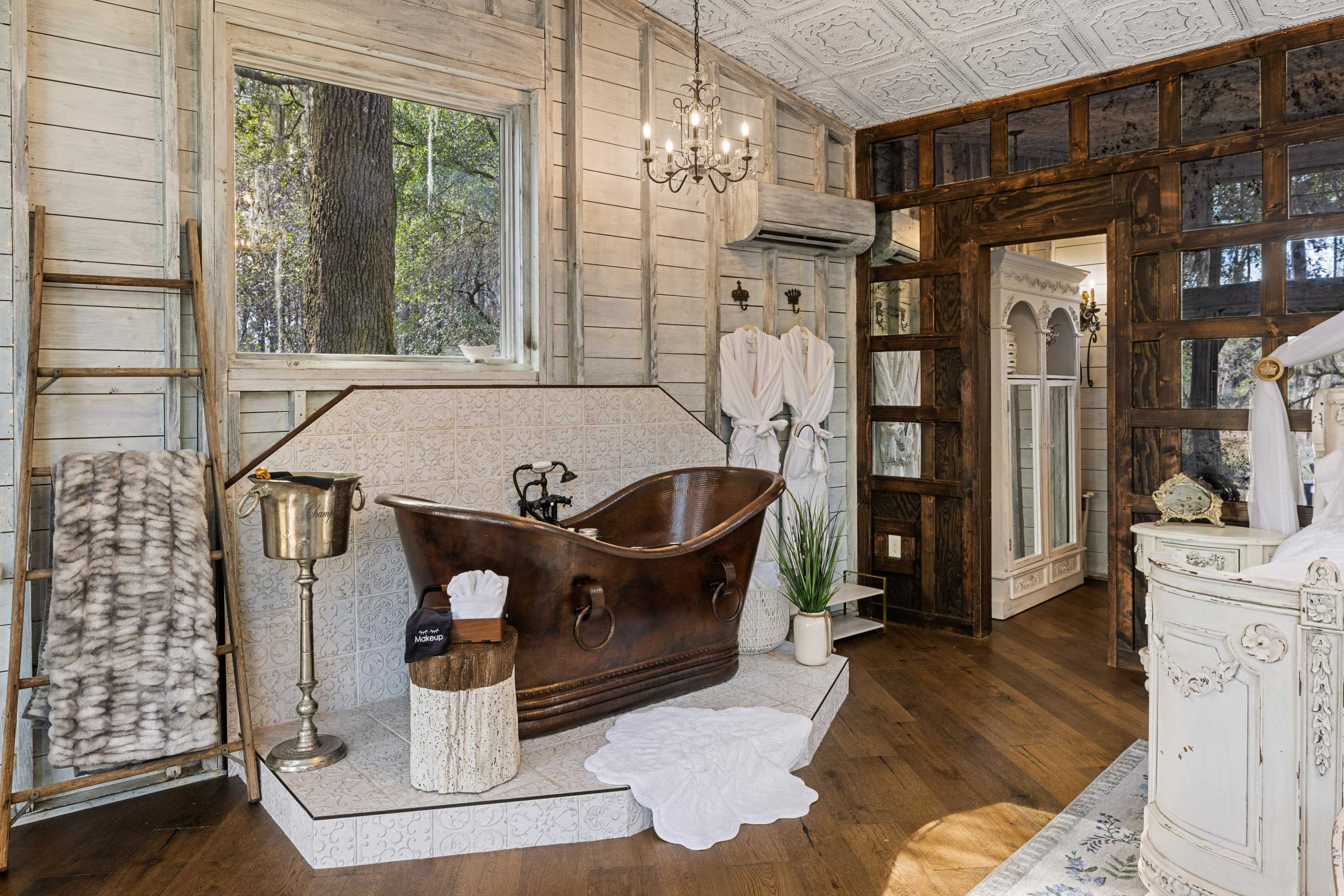 A rustic bathroom featuring a large, freestanding copper bathtub, wooden accents, and a large window with a view of trees outside.