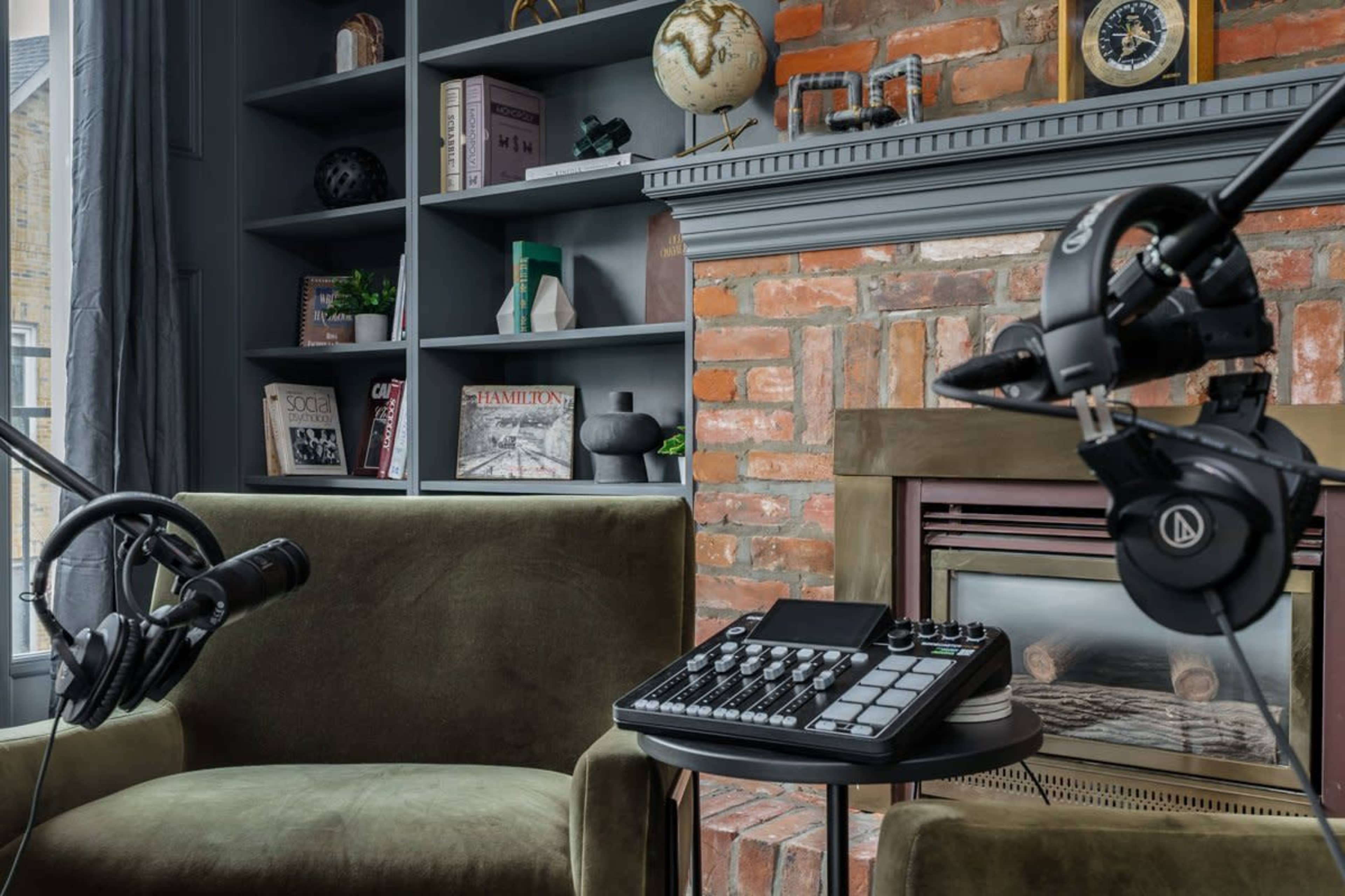 The image shows a cozy room with a brick fireplace, two green chairs, and audio equipment including a microphone and a control pad on a table.