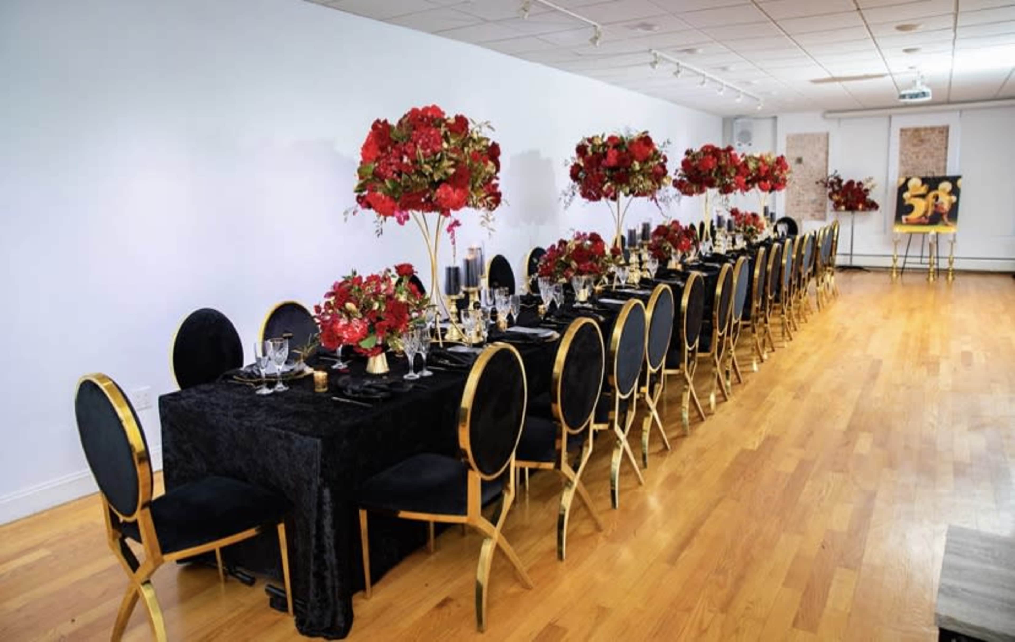 A long banquet table is set with black tablecloths and elegant floral arrangements in red, surrounded by gold-accented chairs.