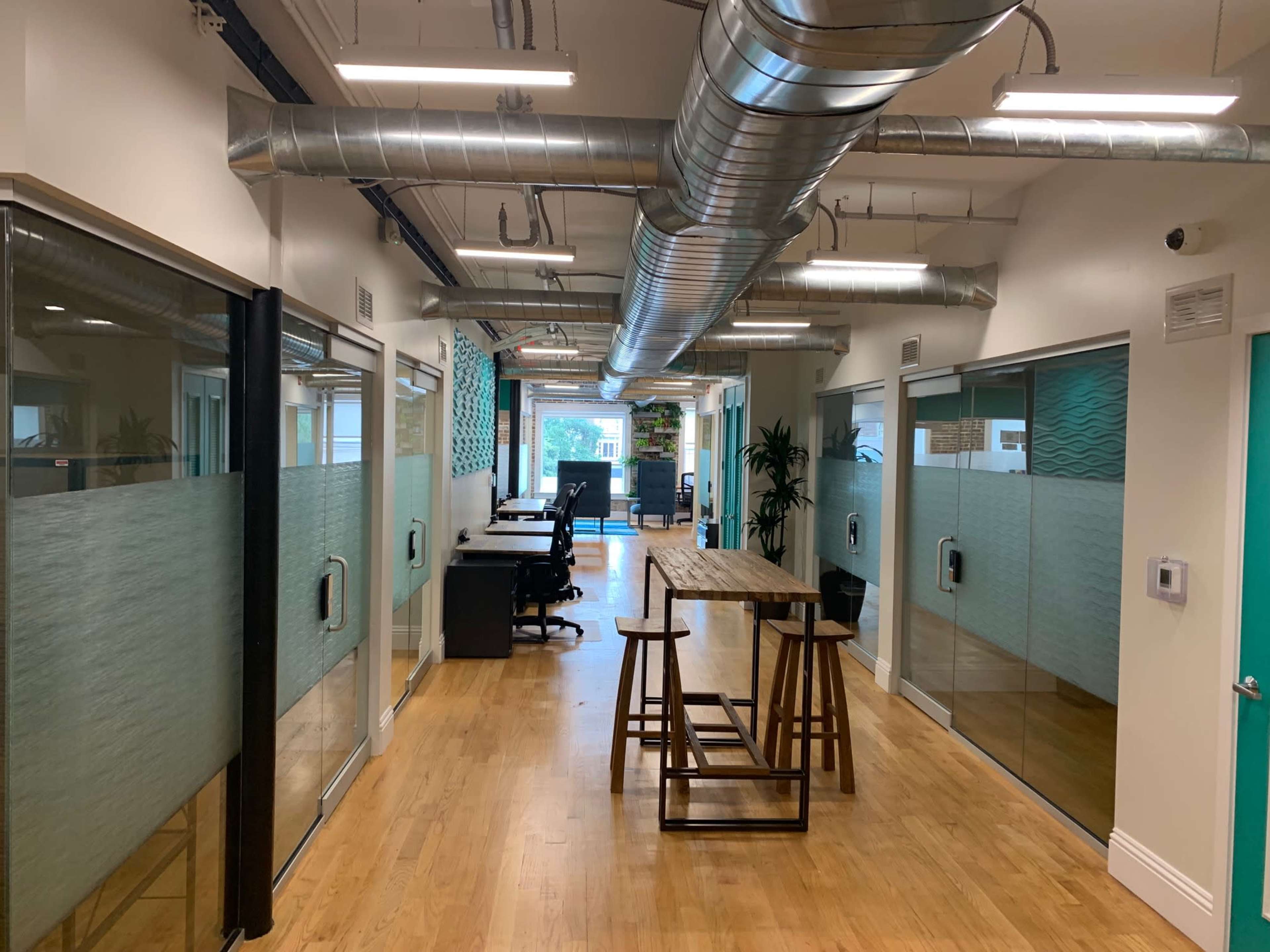 The image shows a modern office corridor with glass meeting rooms on either side and a communal table in the center.