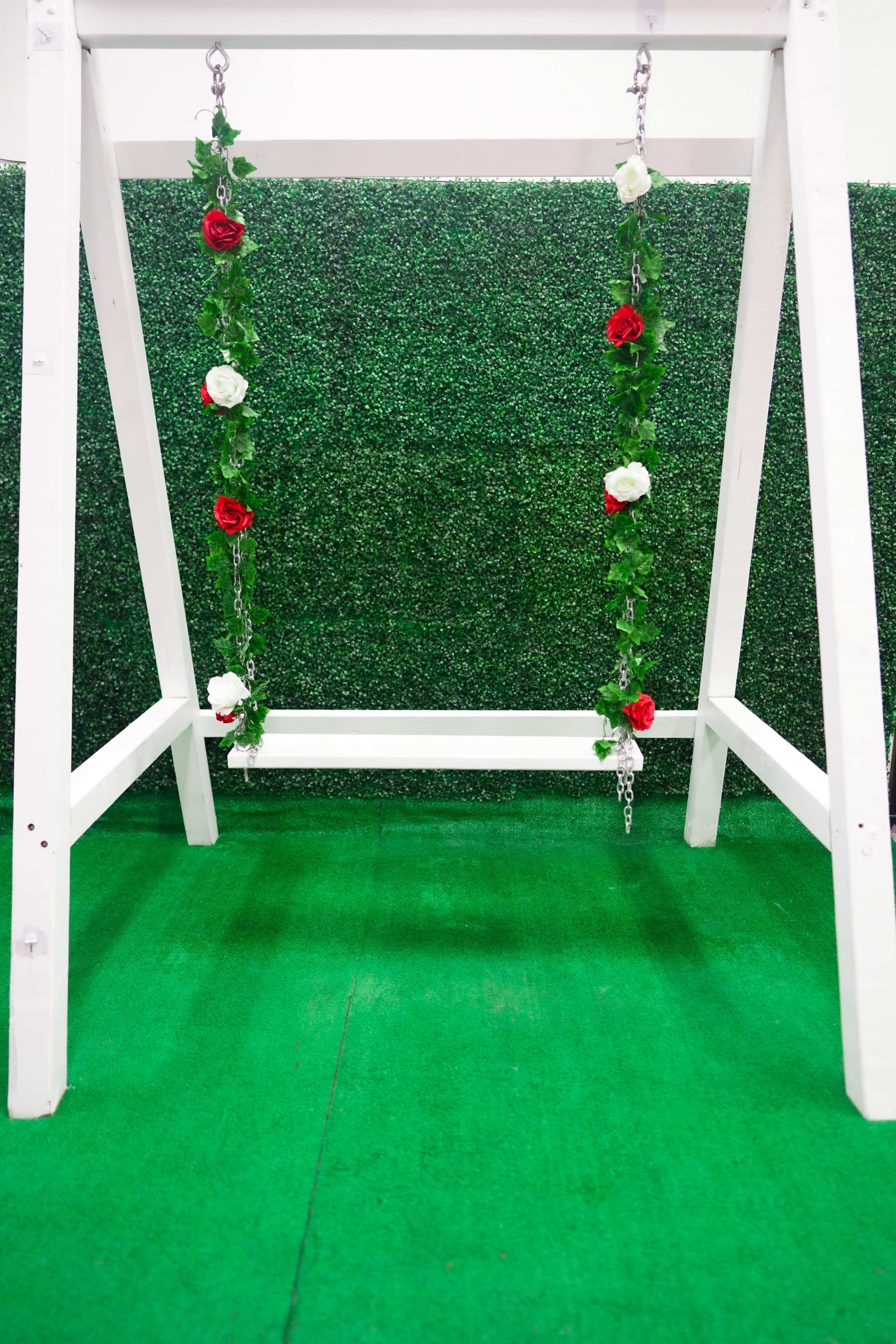 A white swing with floral decorations hangs from a wooden frame against a green hedge backdrop.