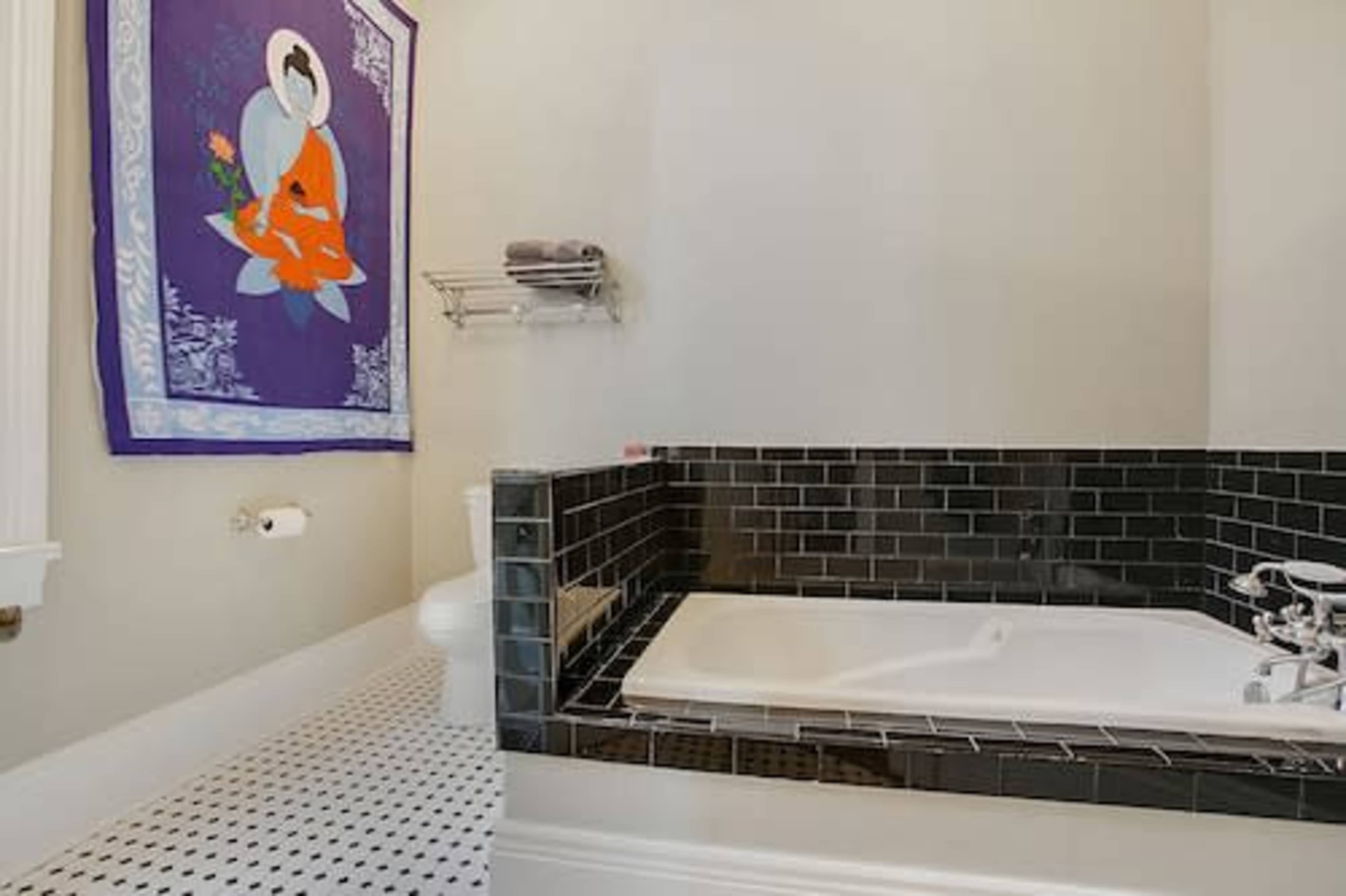 A modern bathroom featuring a black tiled bathtub, a toilet, and a colorful wall hanging.