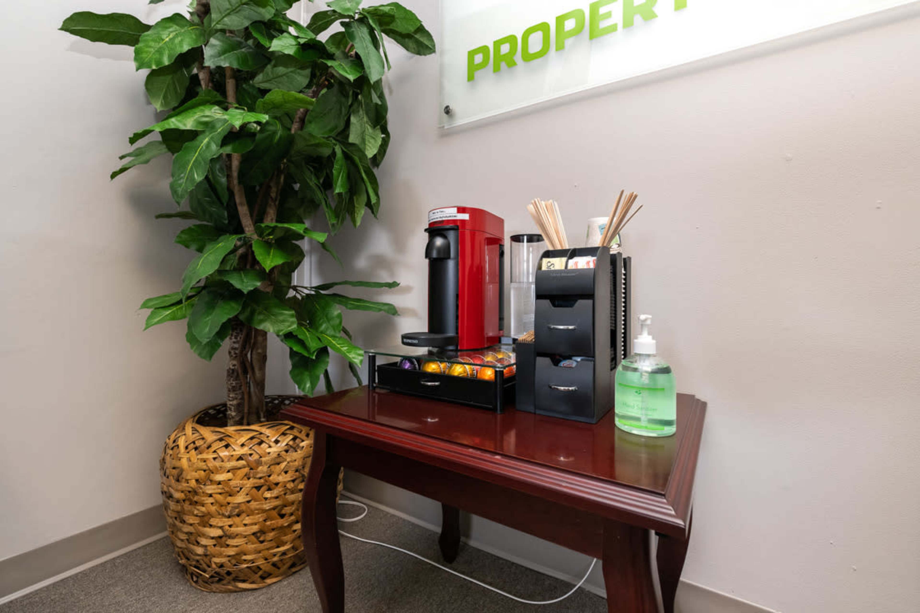 A red coffee maker is placed on a wooden table next to a potted plant and an organizer containing utensils and hand sanitizer.