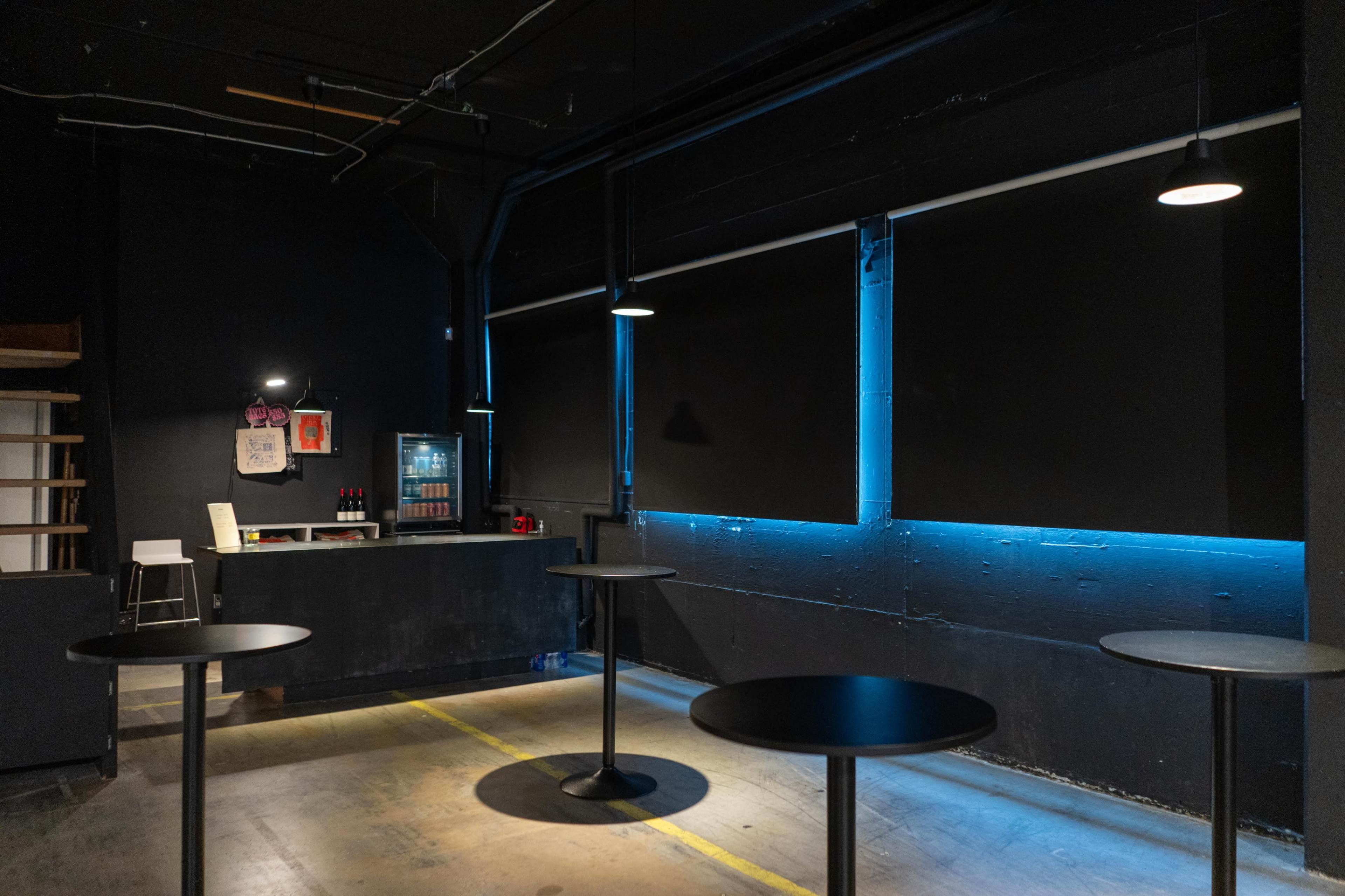 The image shows a dark room with a bar area, black walls, and several round tables illuminated by blue light.