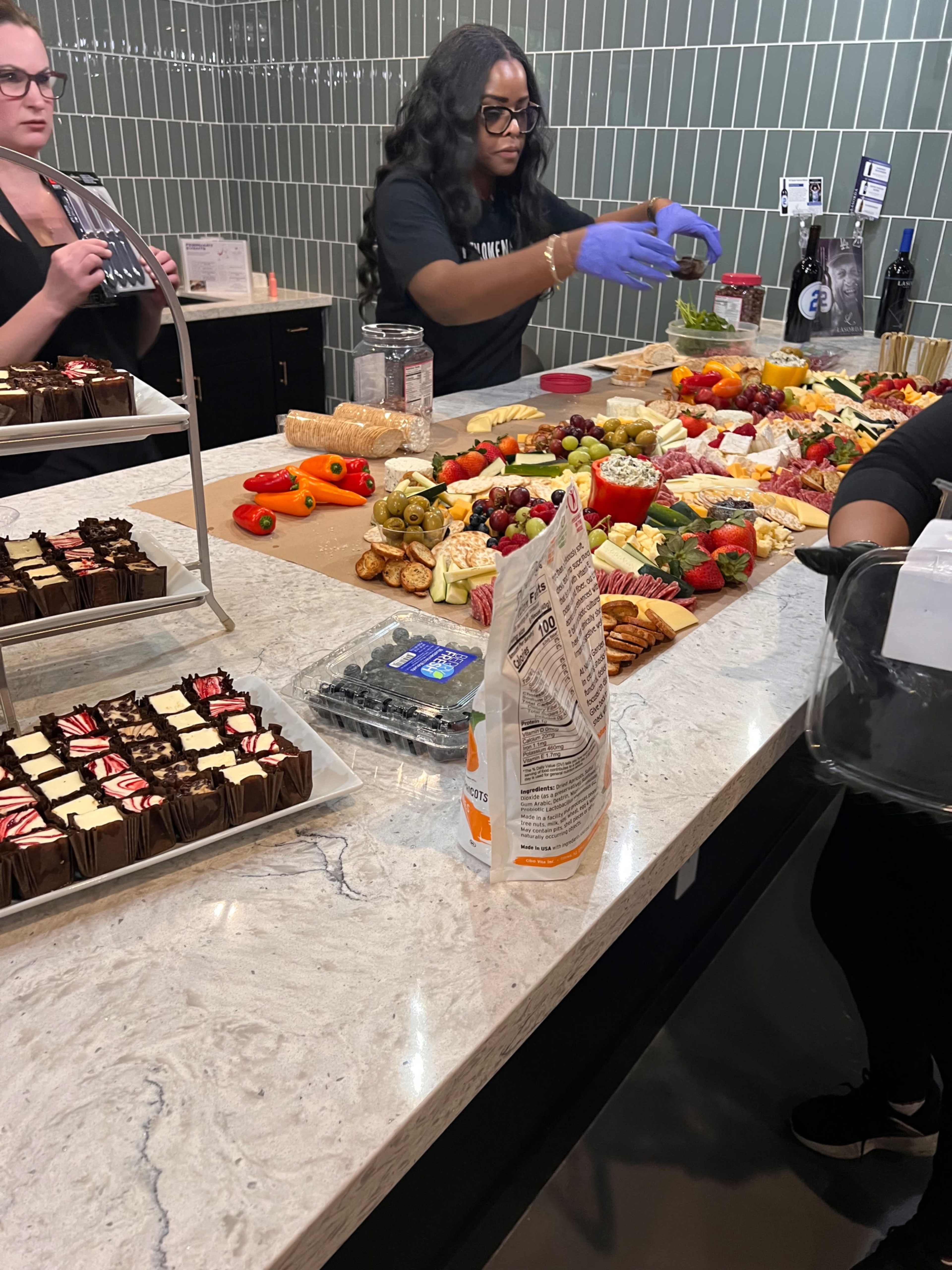 Two people are preparing a large charcuterie board filled with various cheeses, fruits, meats, and snacks on a marble countertop.