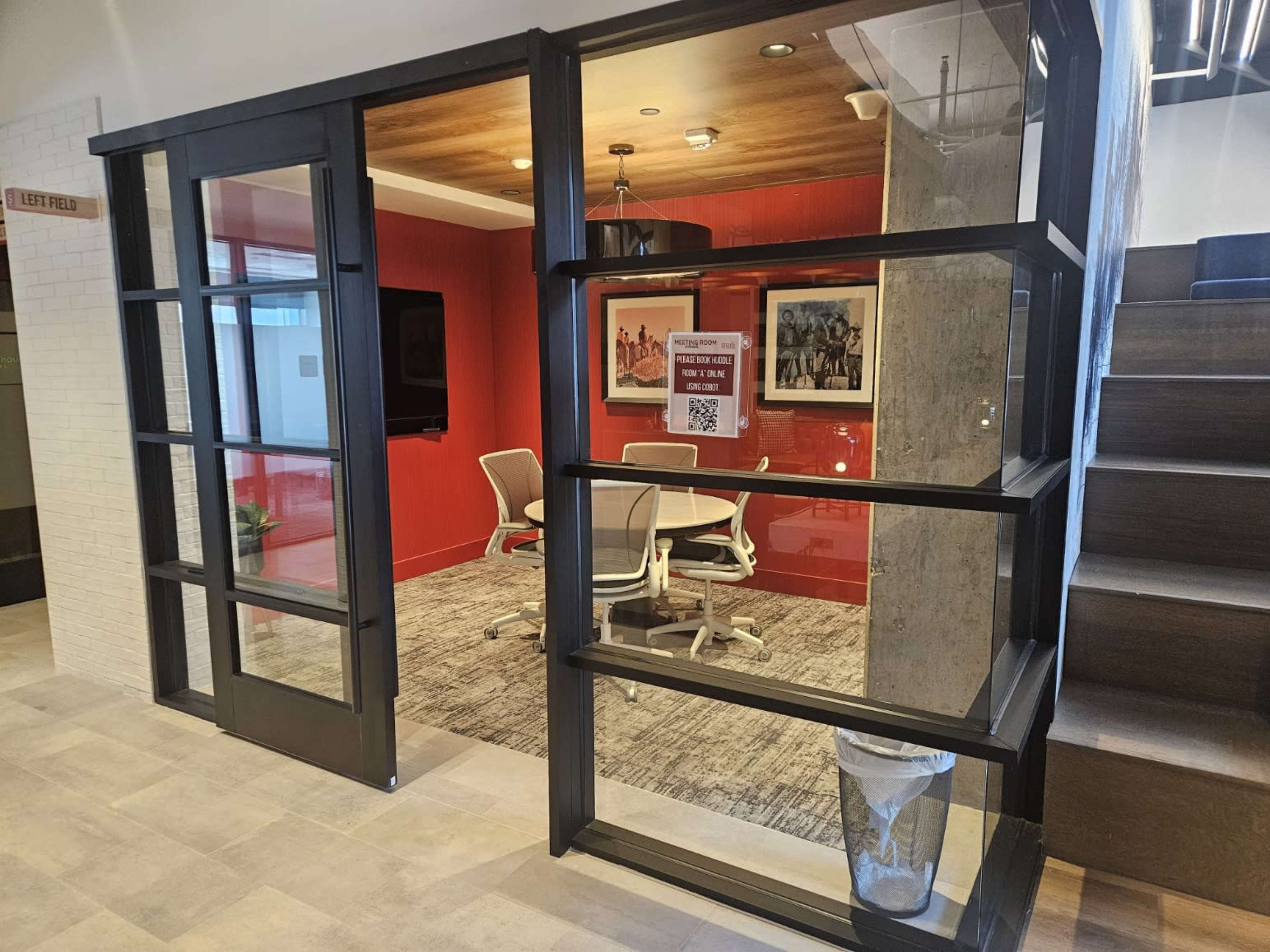 The image shows a modern office space with a glass-fronted conference room featuring a round table and chairs, surrounded by a red accent wall.