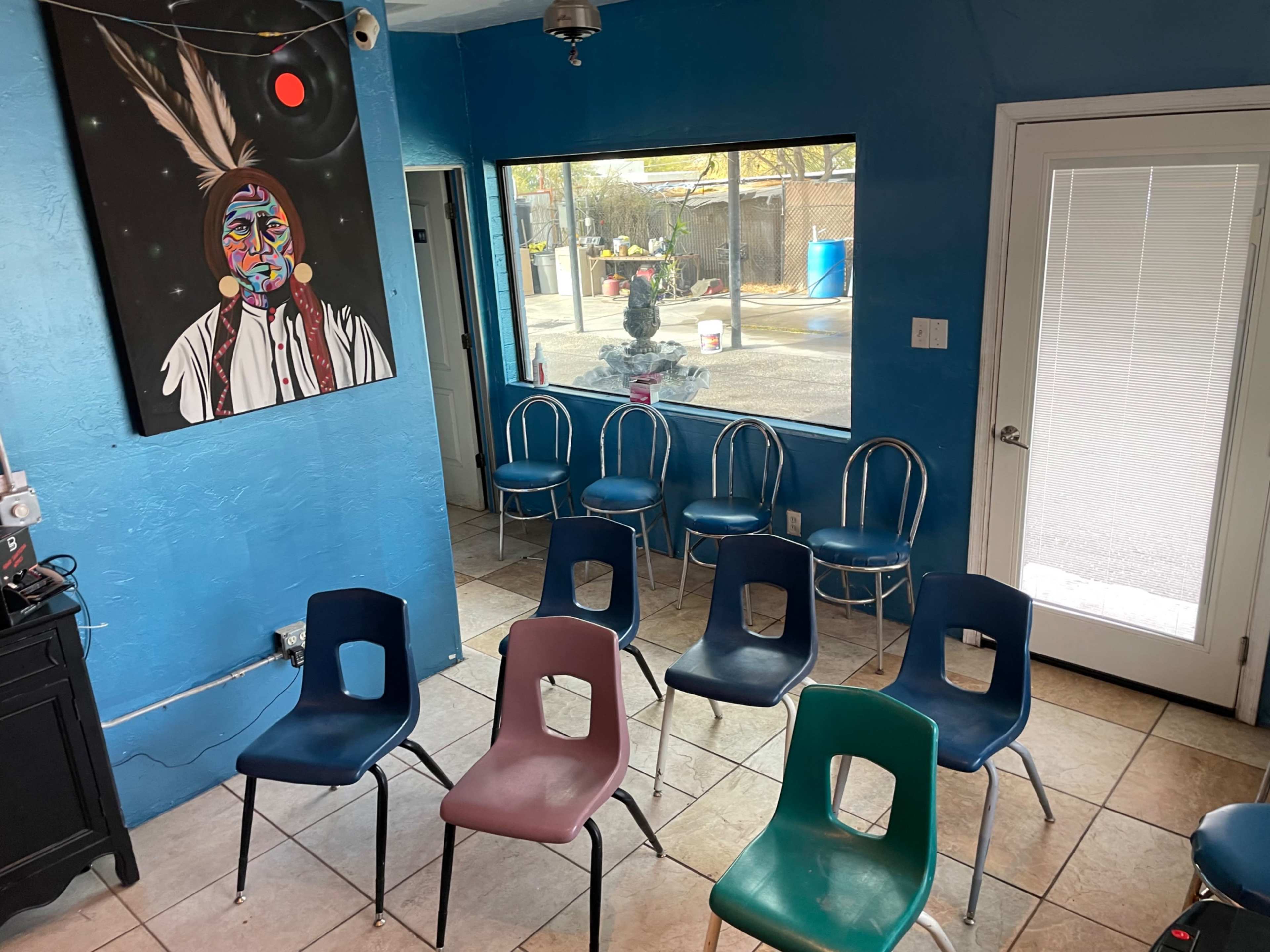 The image shows a blue-walled room with a painting on one wall, several metal and plastic chairs arranged in a semicircle, and a large window facing an outdoor area.