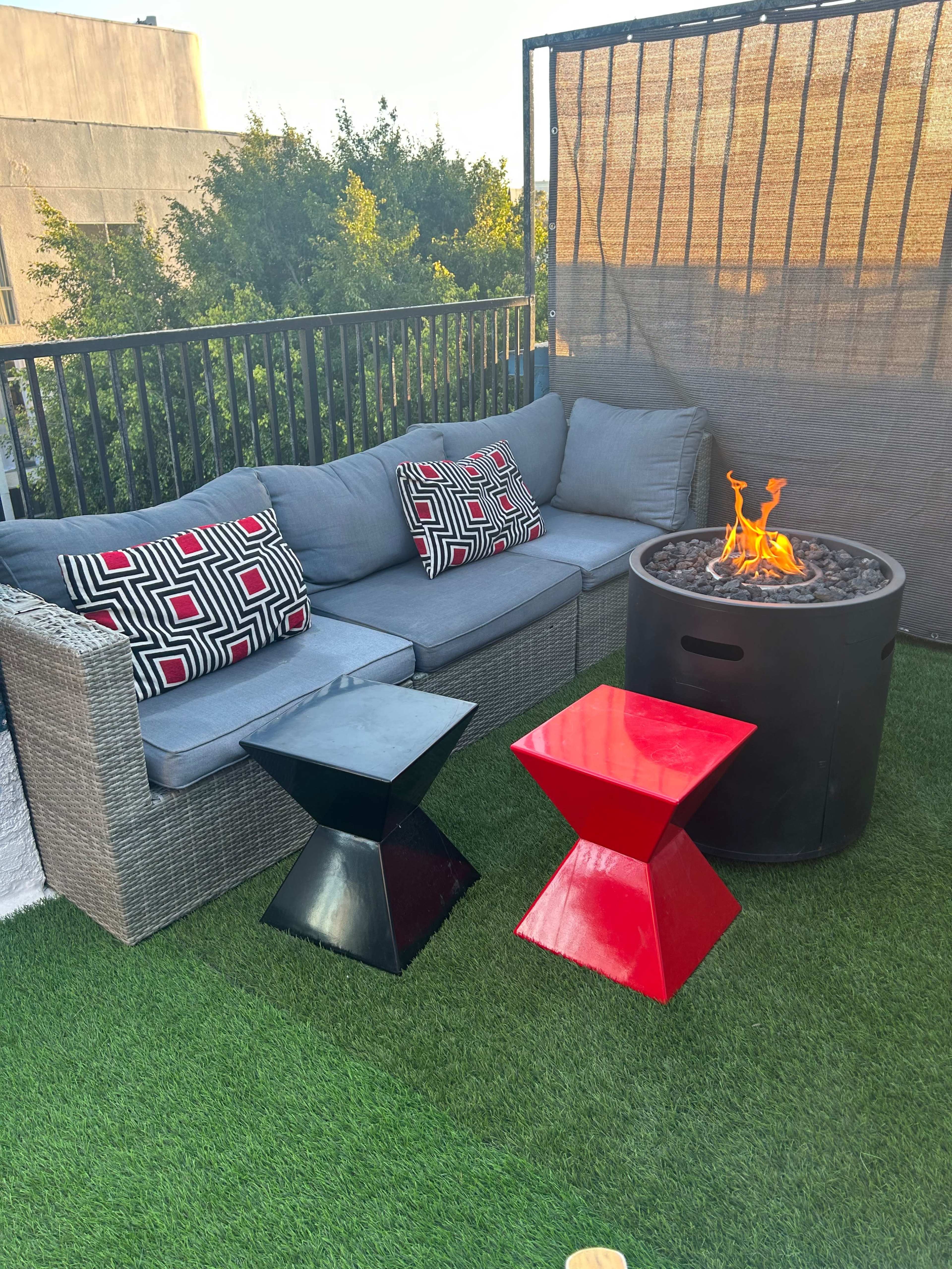 A patio area features a grey sectional sofa with patterned cushions, two colorful side tables, and a circular fire pit surrounded by artificial grass.