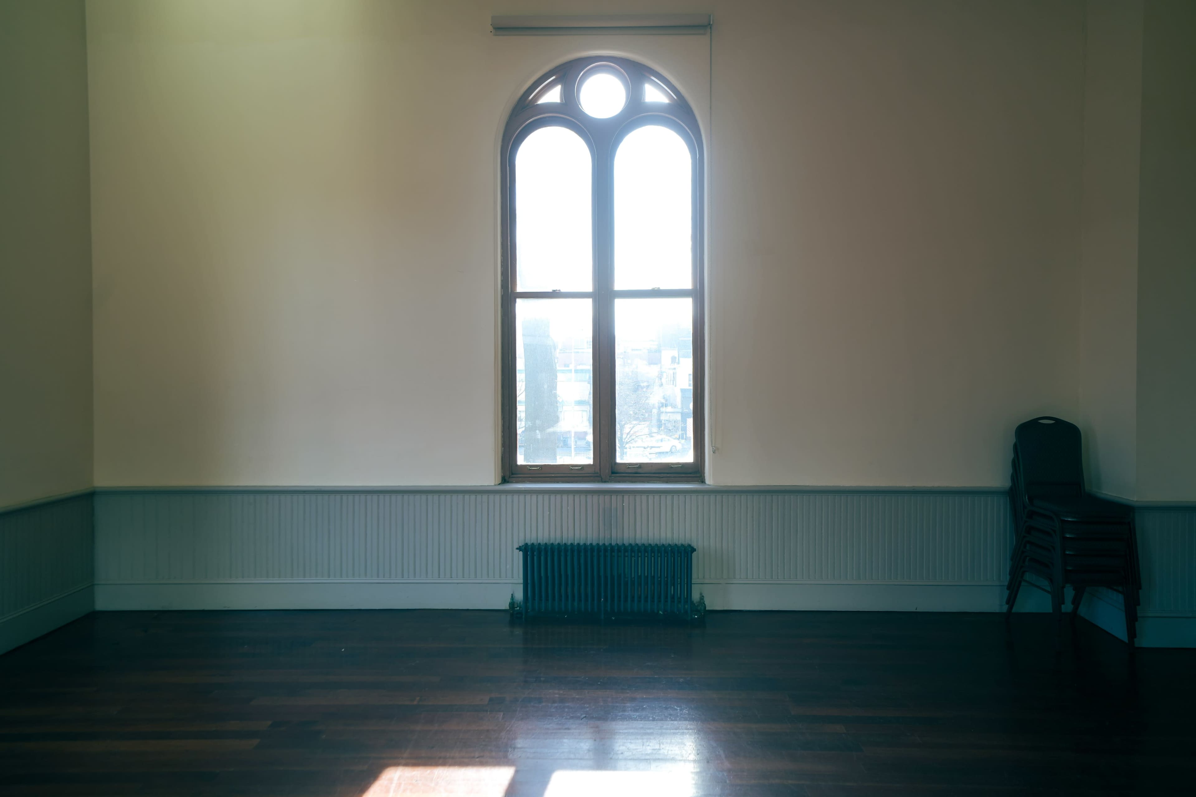 A large window with an arched top is positioned on a bare wall in a spacious, empty room with wooden flooring and a radiator beneath the window.