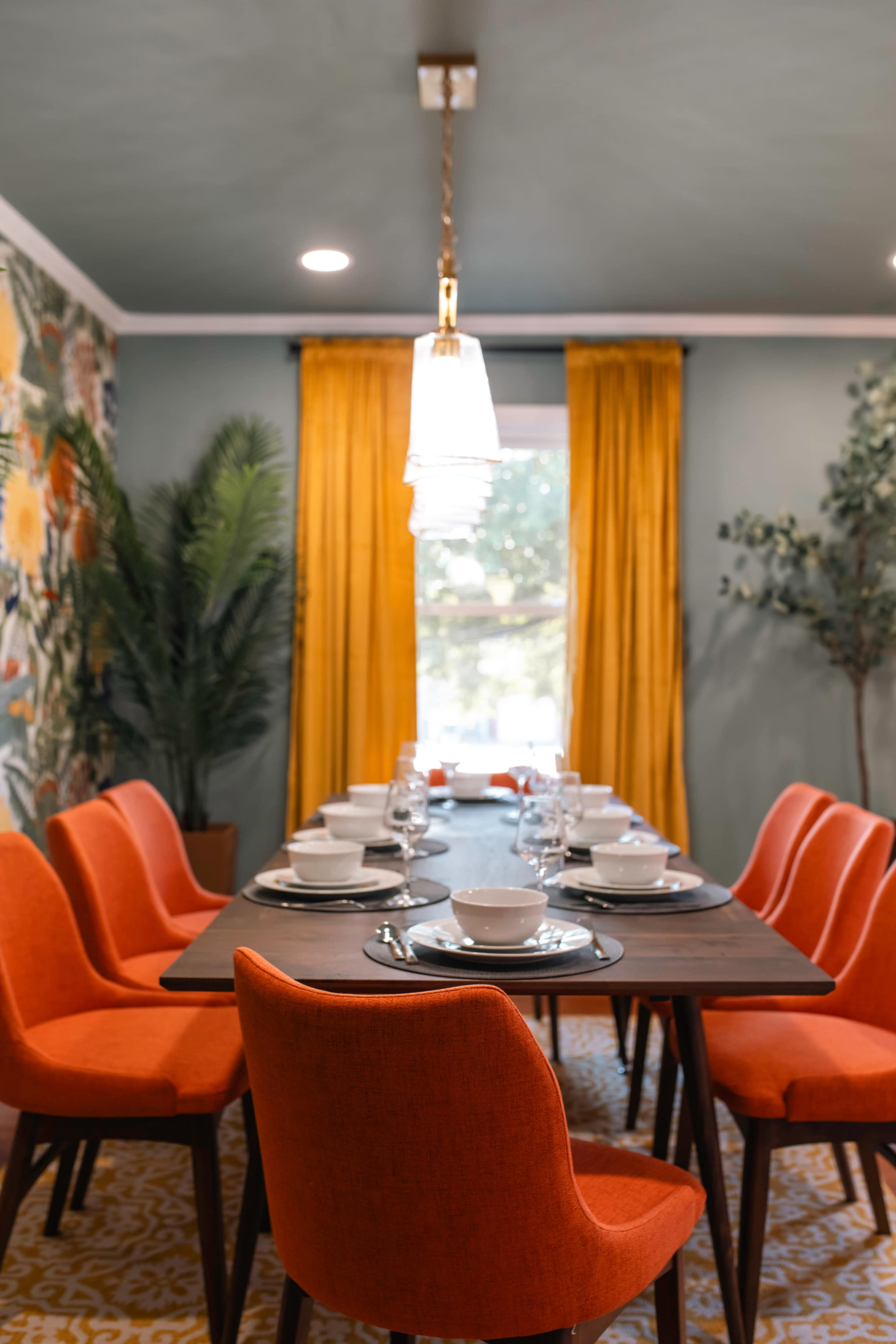 A modern dining room features a long table set with dishware and surrounded by bright red chairs, with floral wallpaper and yellow curtains framing a window.