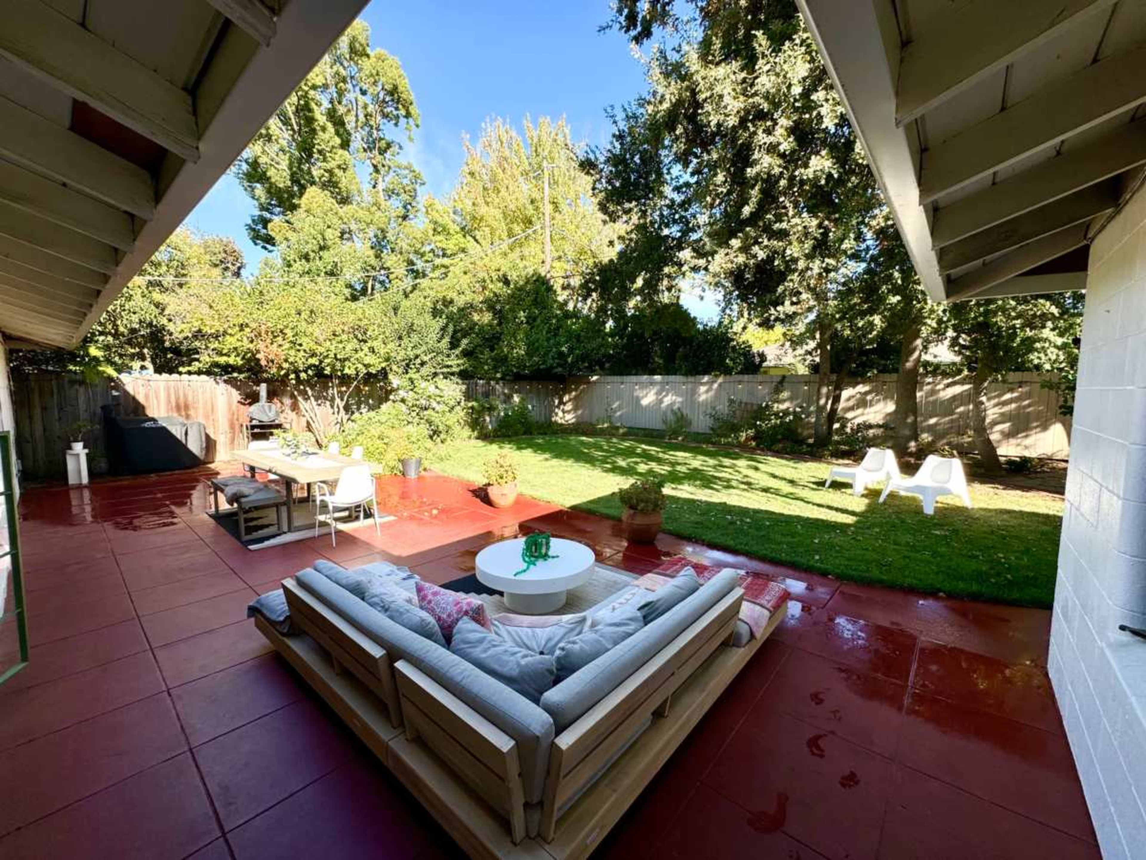 A spacious backyard features a patio with outdoor seating, a dining area, and a verdant lawn surrounded by trees.