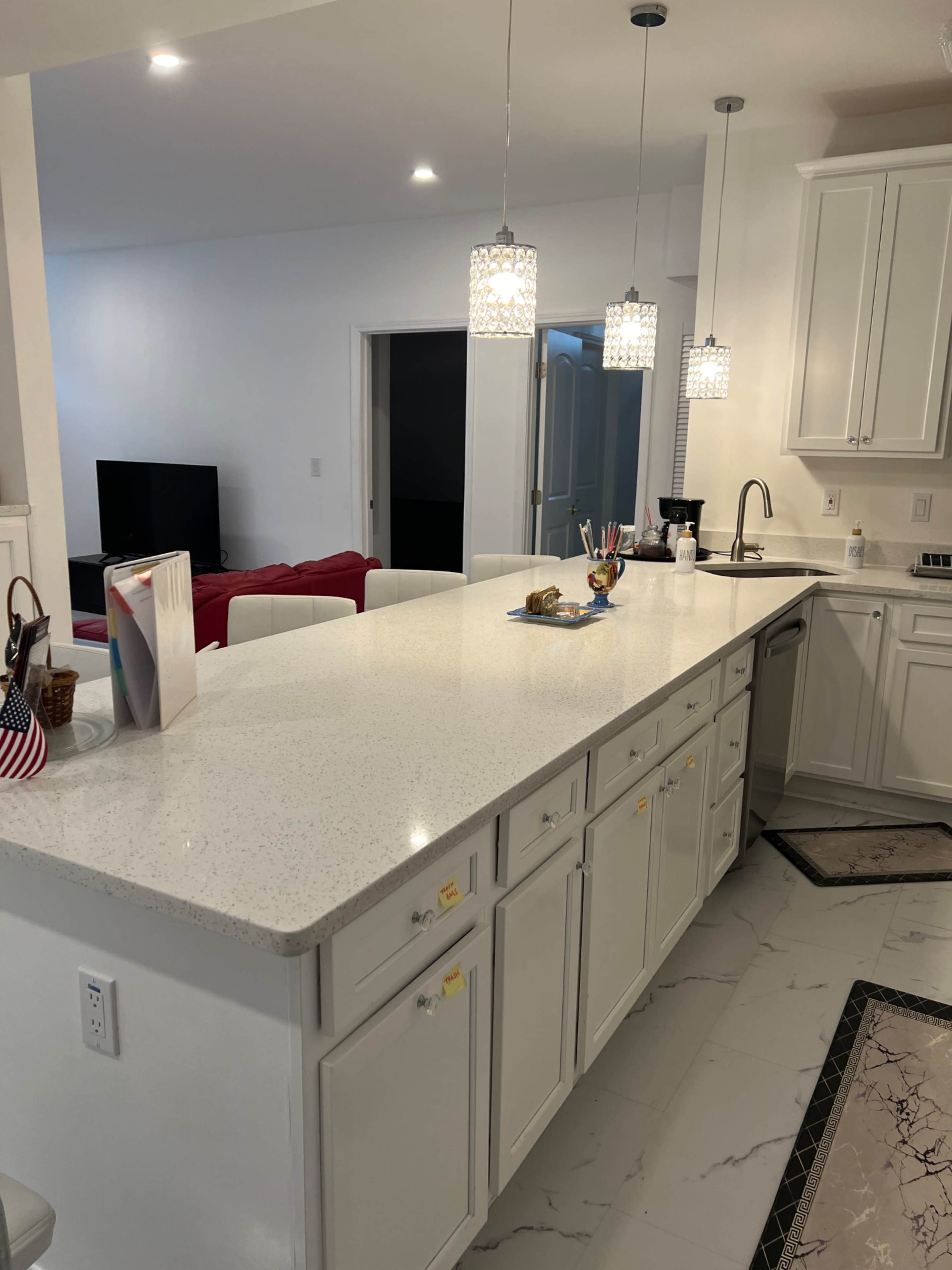The image shows a modern kitchen featuring a white countertop, pendant lights, and a view into a living area with a red couch.
