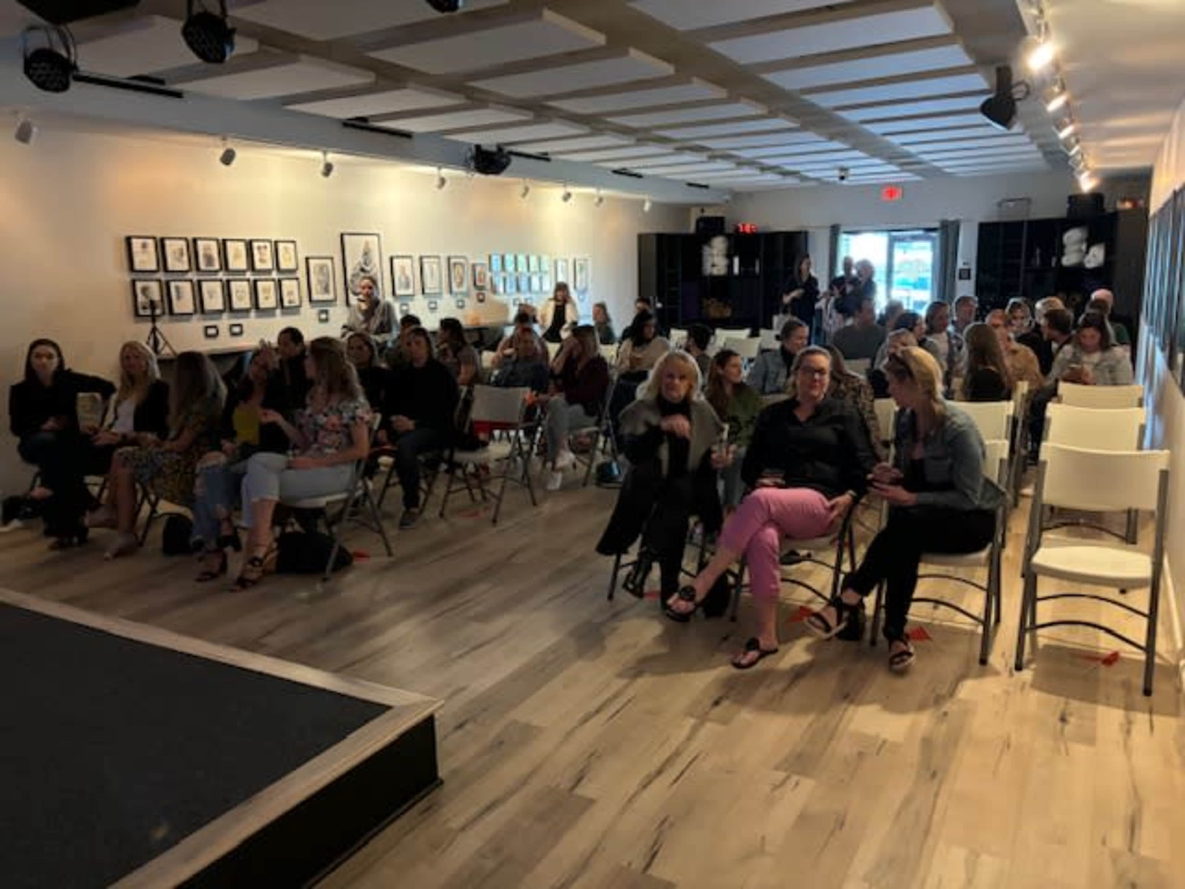 A seated audience fills a well-lit room adorned with framed artwork, waiting for an event to begin.