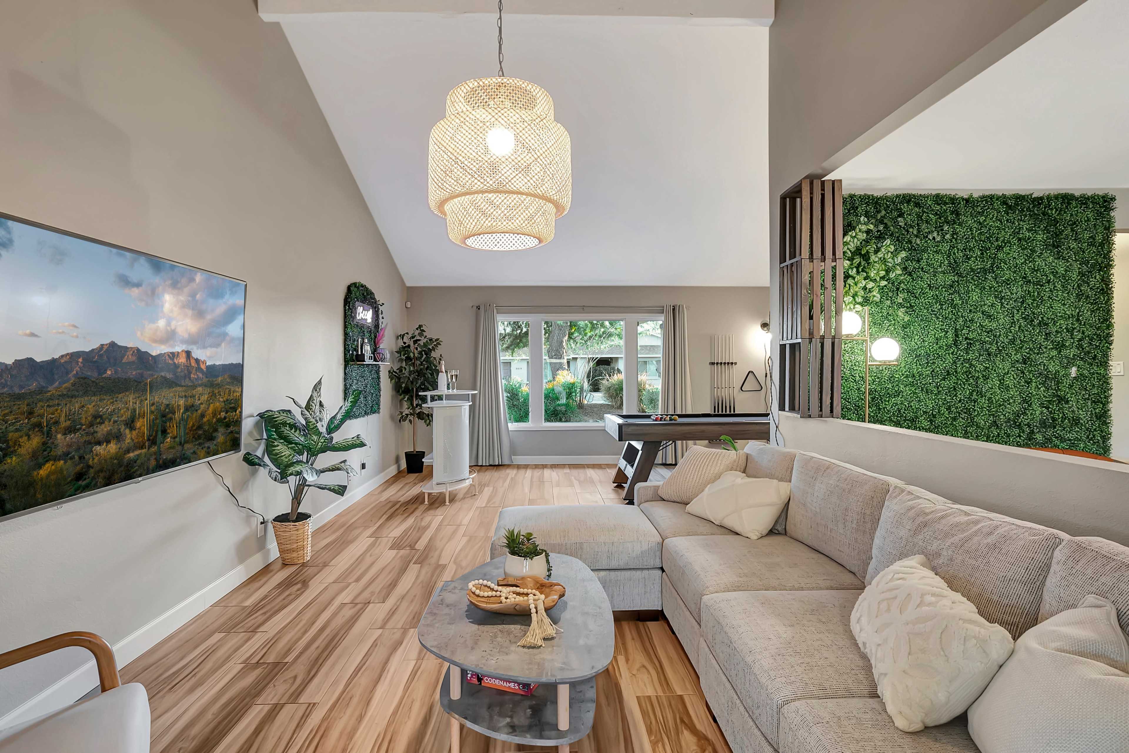 A modern living room featuring a light-colored sectional sofa, a large wall-mounted TV, and a green vertical garden beside a pool table.