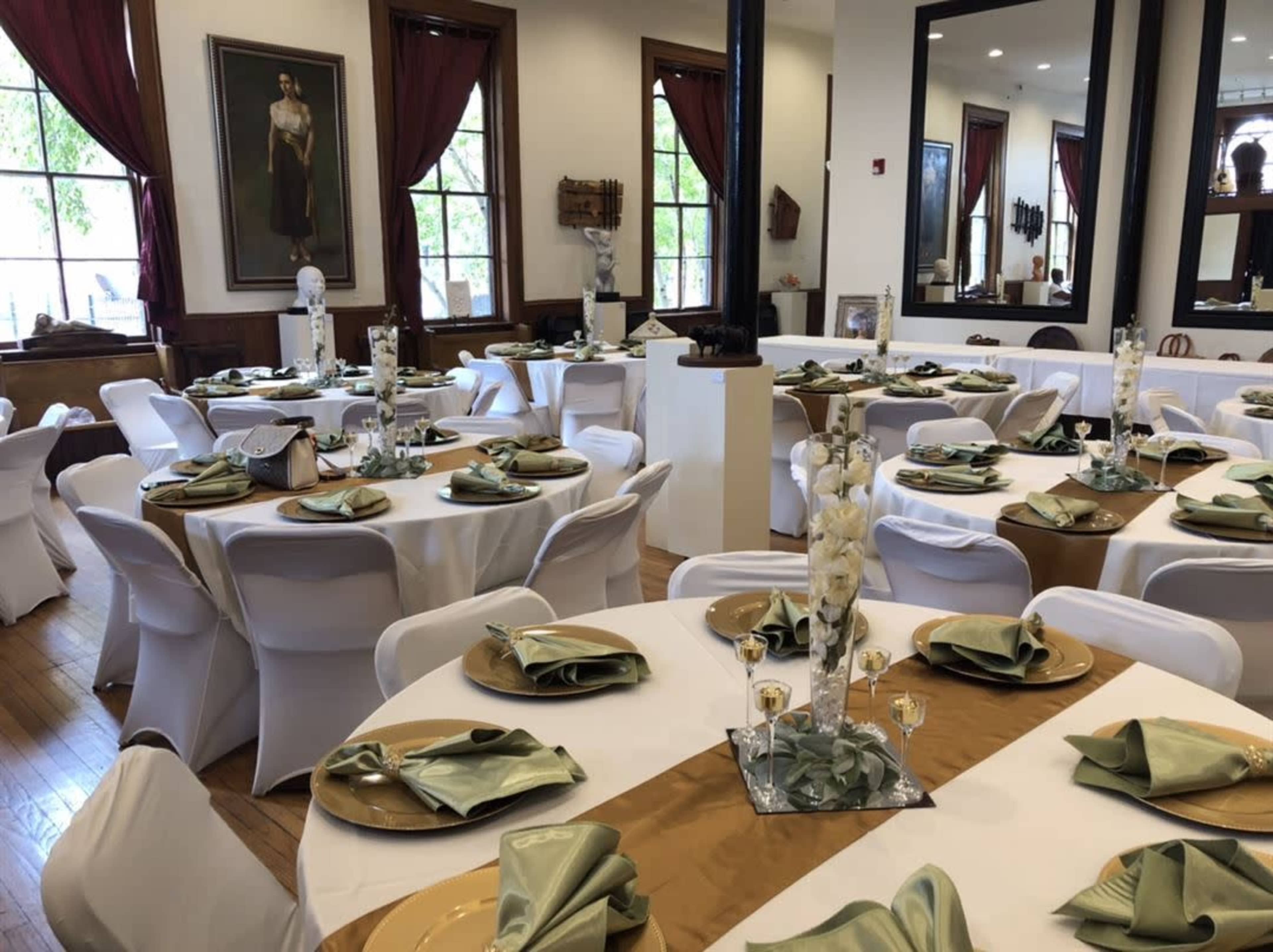 The image shows a banquet hall set with round tables that are covered with white tablecloths and adorned with green napkins and gold plates.