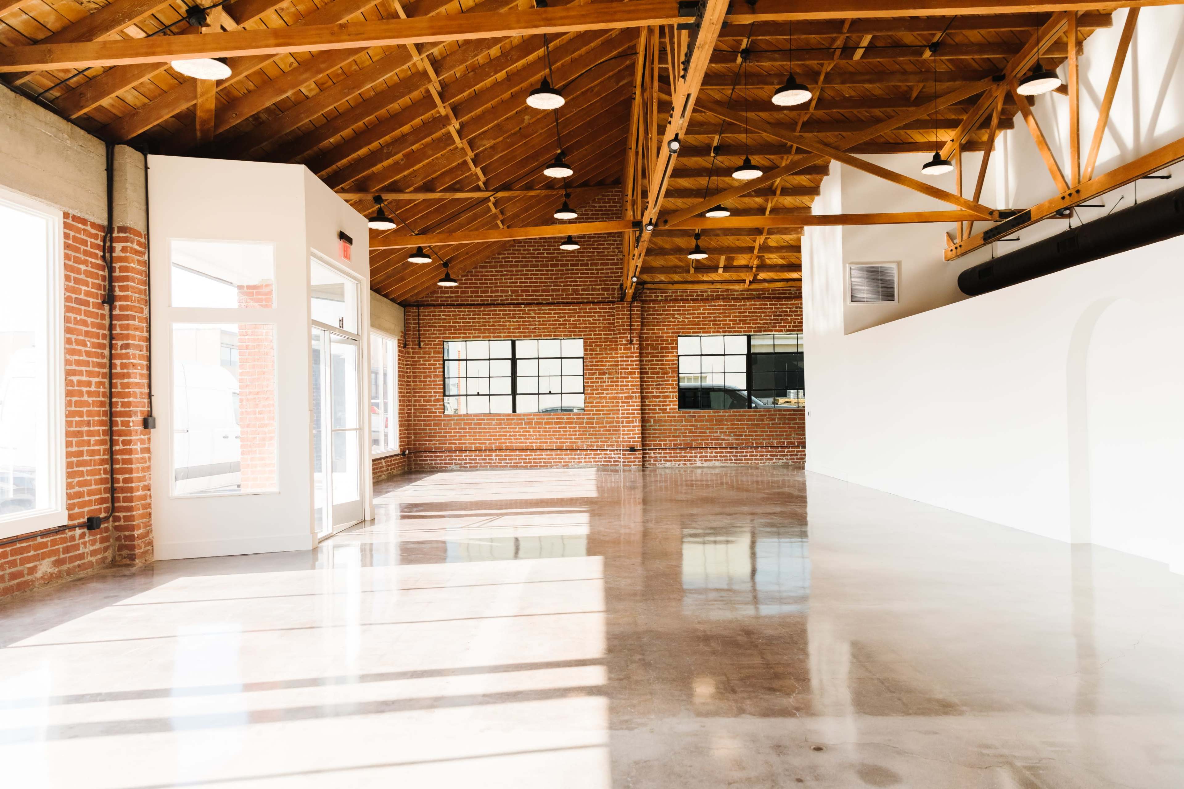 A spacious interior of a commercial building with a high wooden ceiling, exposed brick walls, large windows, and polished concrete floors.